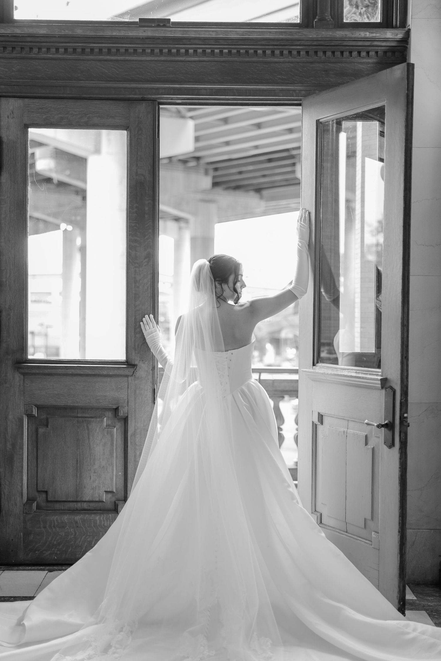 Bride in flowing white gown and veil opens vintage wooden doors in black and white photograph.