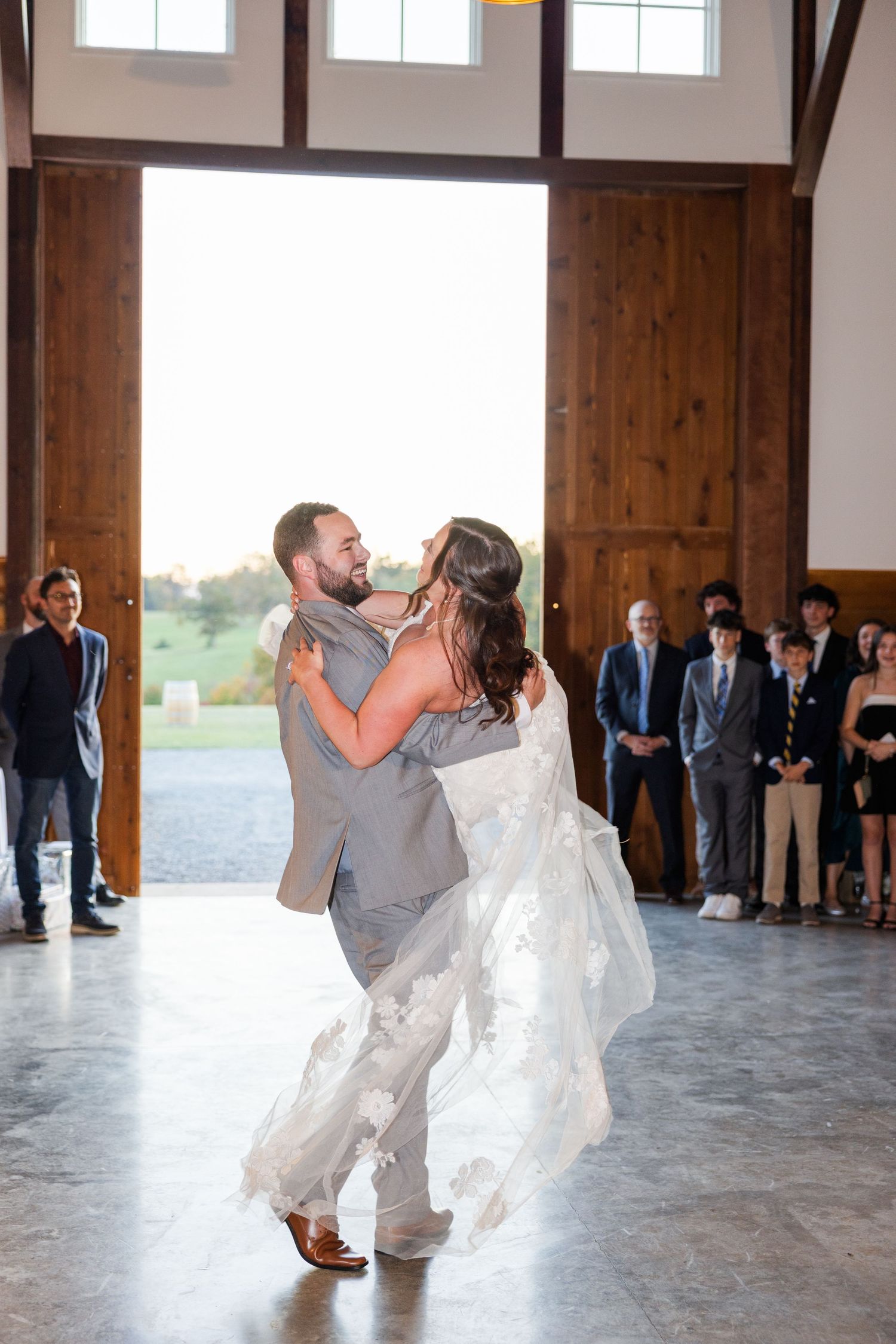 A couple performs their first dance in a spacious rustic venue with wooden beams and large windows.