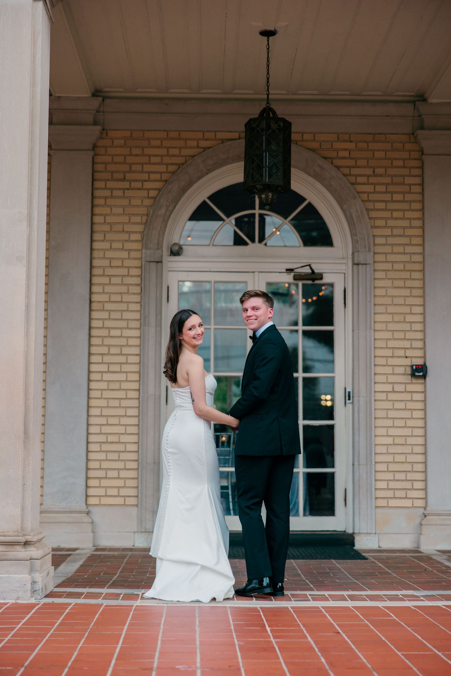 Wedding couple on brick porch with arched windows and classical architecture.