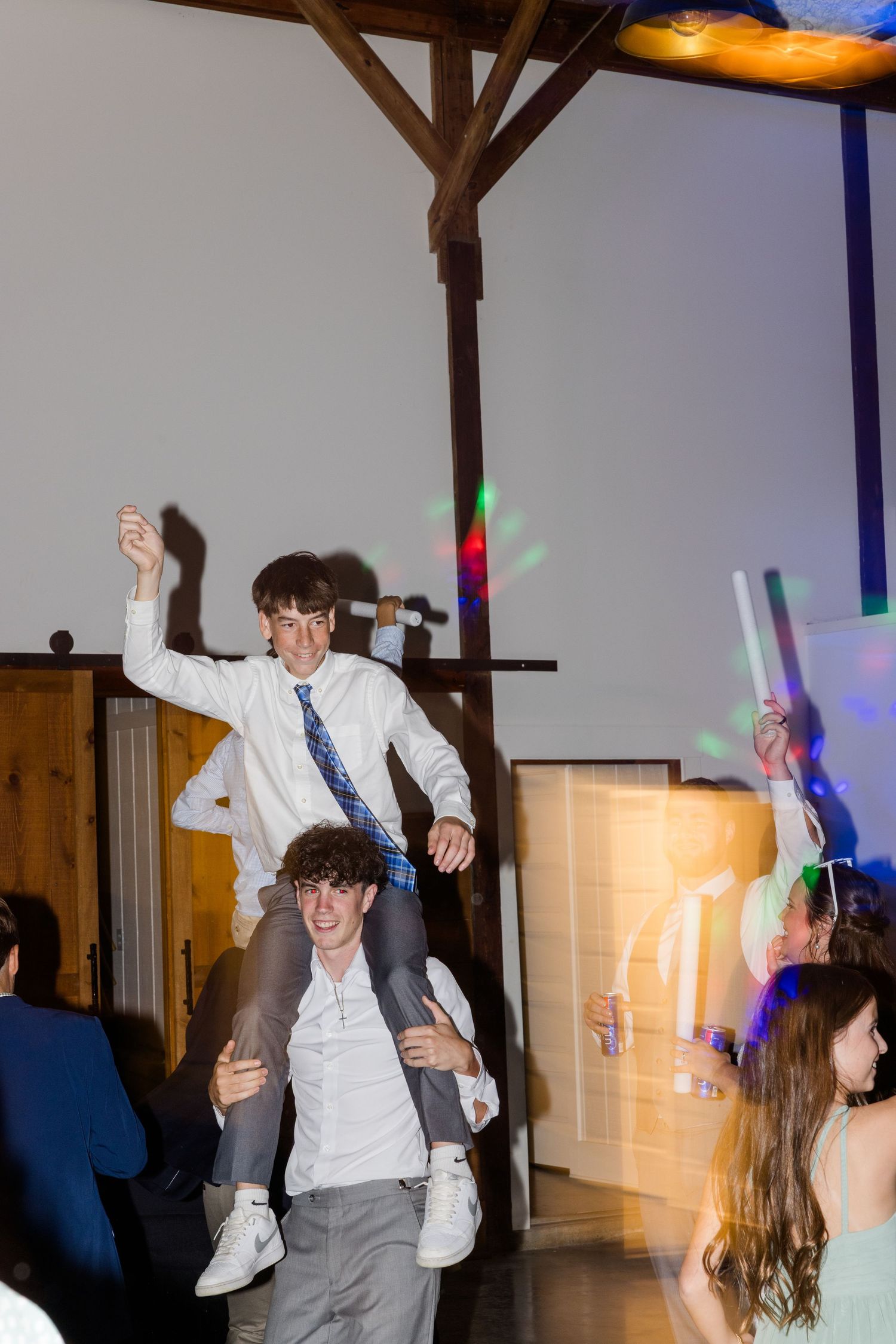 Two young men having fun at a party with one riding on the other's shoulders while dancing under colorful lights.