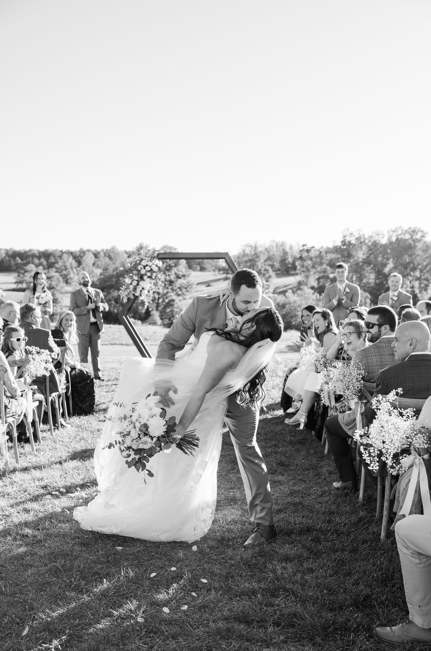 Black and white photo of wedding guests watching ceremony from white chairs on outdoor lawn.