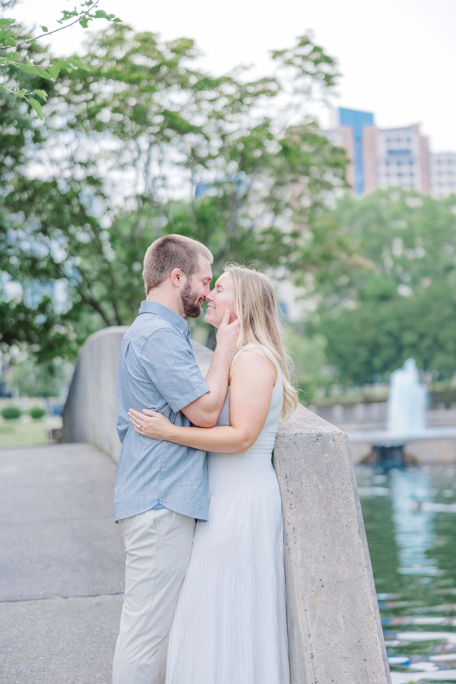 A couple embraces tenderly along a waterfront path with urban buildings and trees in the background.