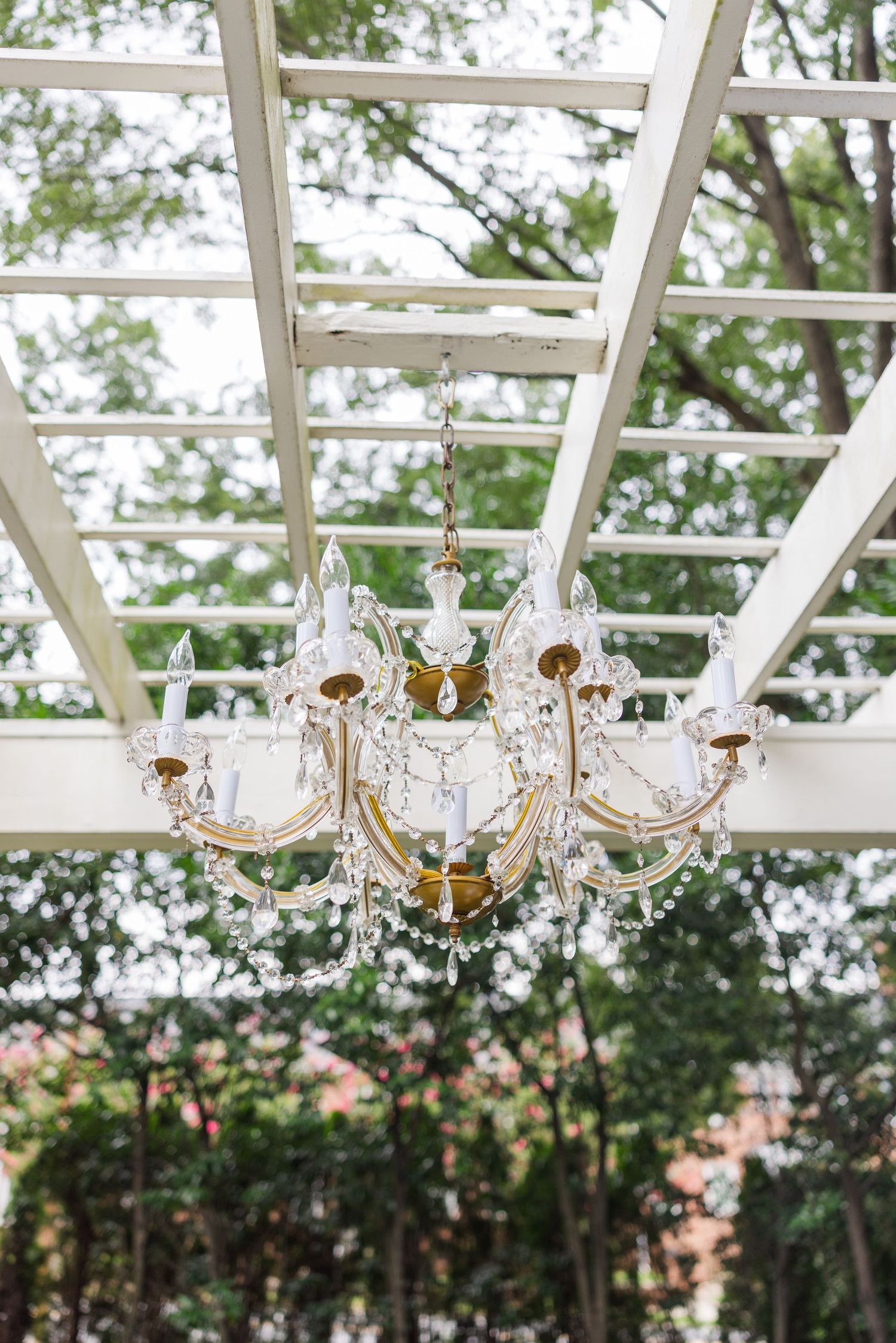 Crystal chandeliers hang from a white pergola structure with lush greenery visible in the background.