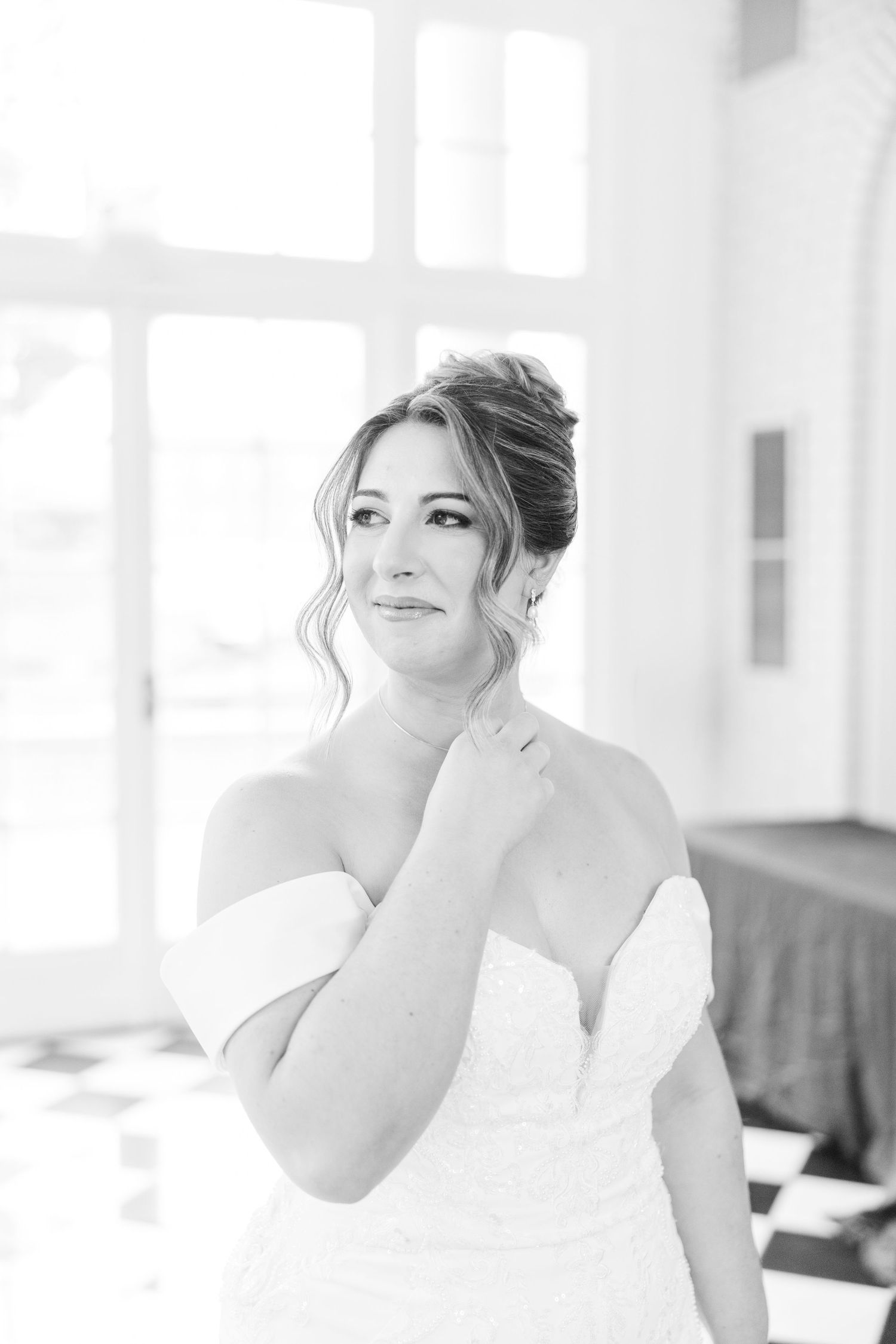 A bride in a white off-shoulder gown poses elegantly in soft window light.
