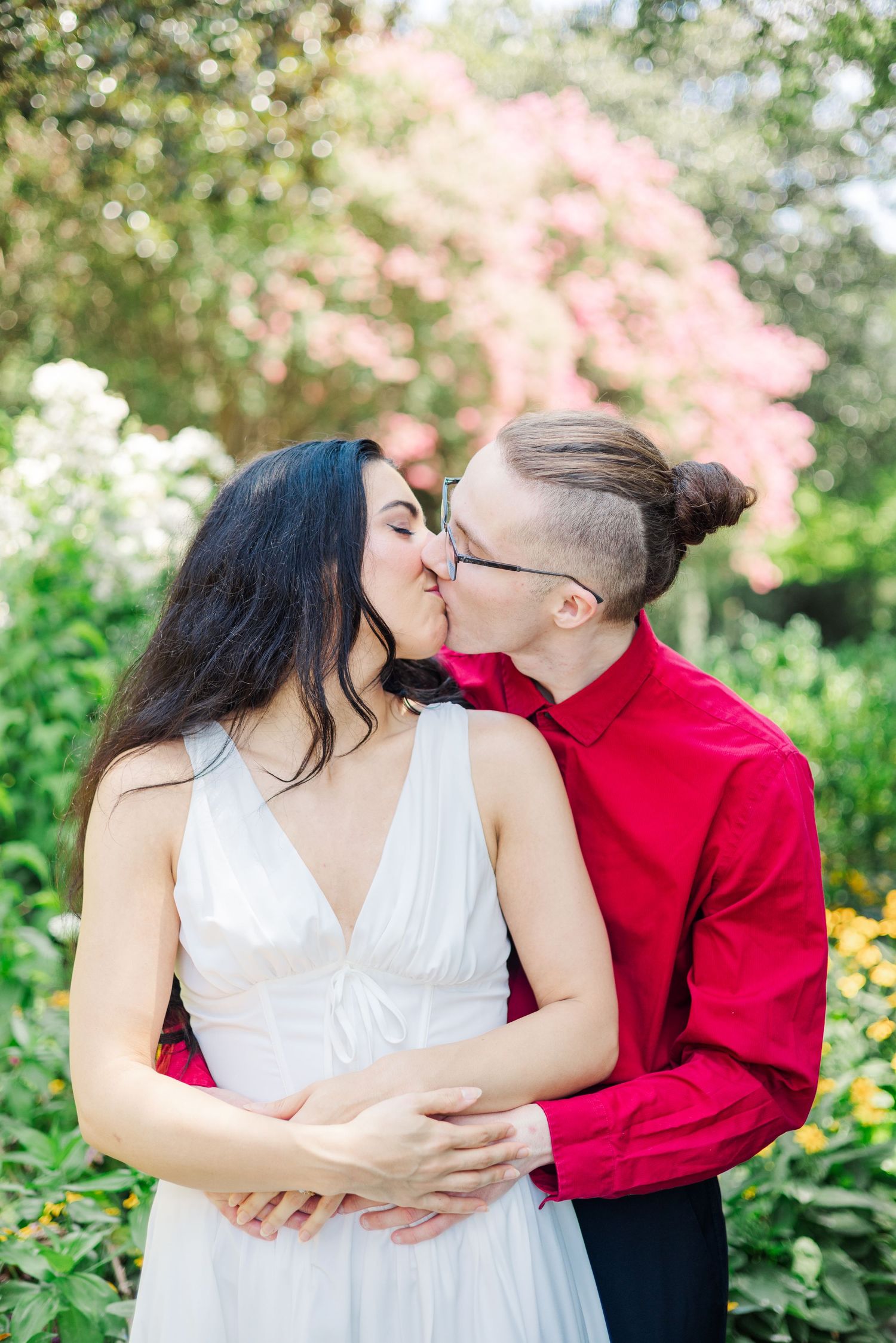 A couple shares an intimate moment in a garden surrounded by pink blossoms and yellow flowers.