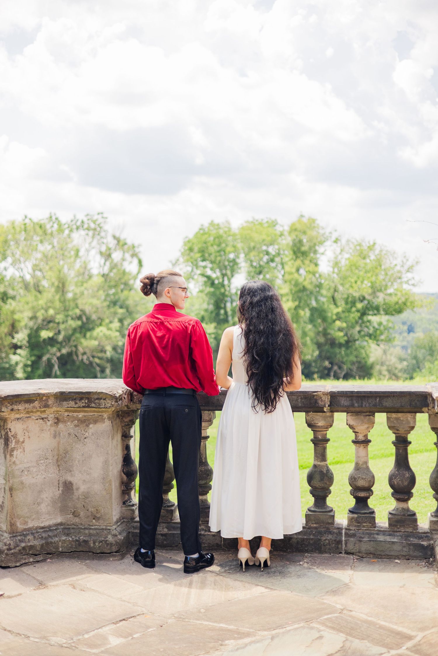 A couple stands at a stone balustrade overlooking a scenic landscape on a sunny day.