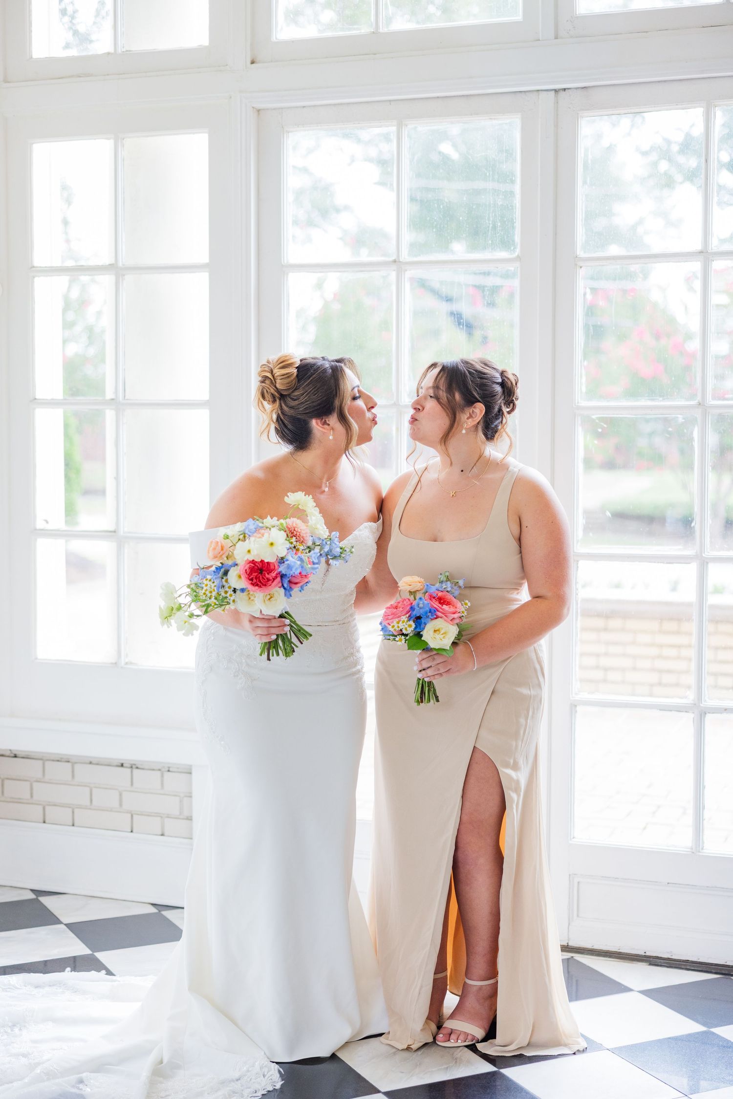 Two people in cream-colored dresses hold colorful bouquets in a bright white room with checkerboard flooring.