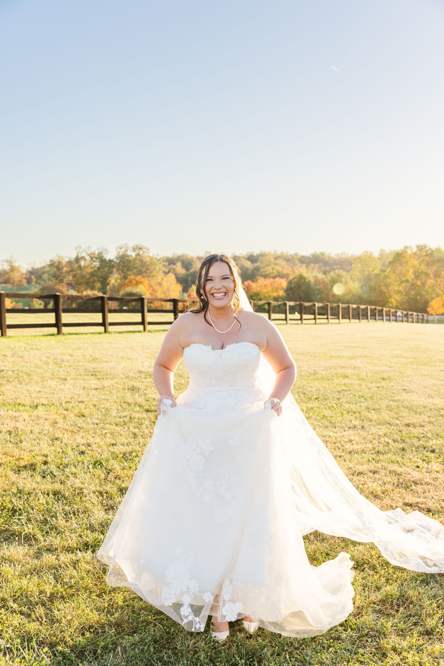 Bride in a white ballgown poses in a sunny field with wooden fences in the background.