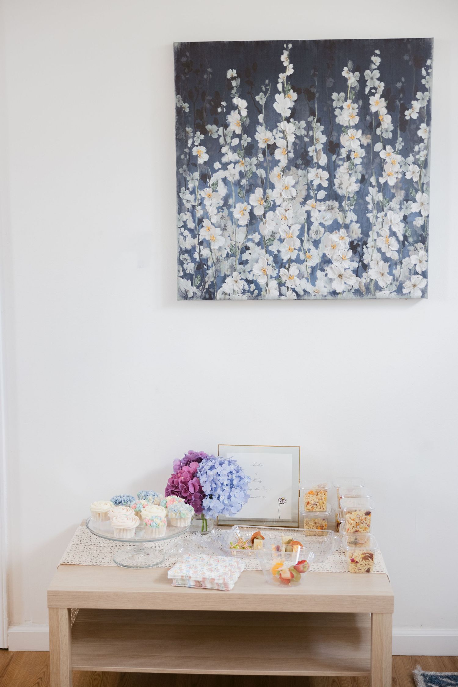 Wedding reception table setup with floral artwork and decorative items against a white wall.