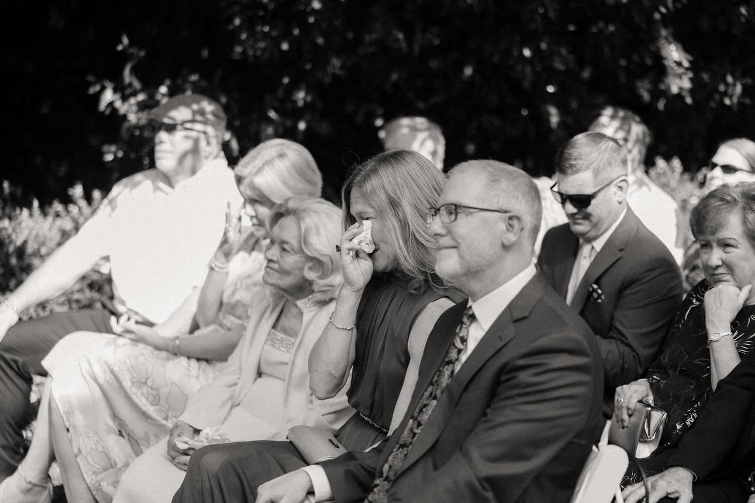 A black and white photograph shows spectators sitting in chairs outdoors at an event in formal attire.