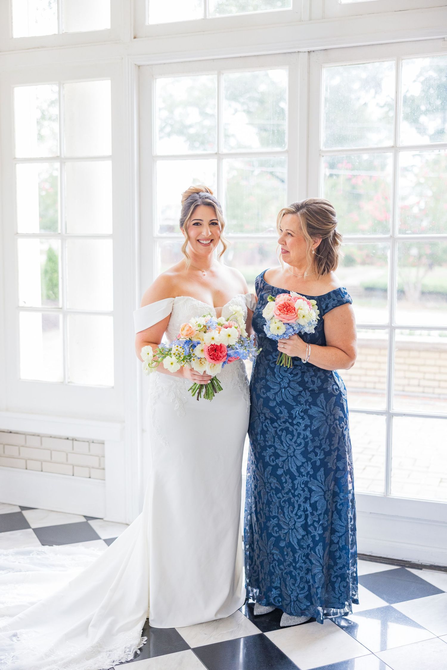 Two women in a bright sunlit room share a joyful moment, one wearing a navy lace dress and one in white.