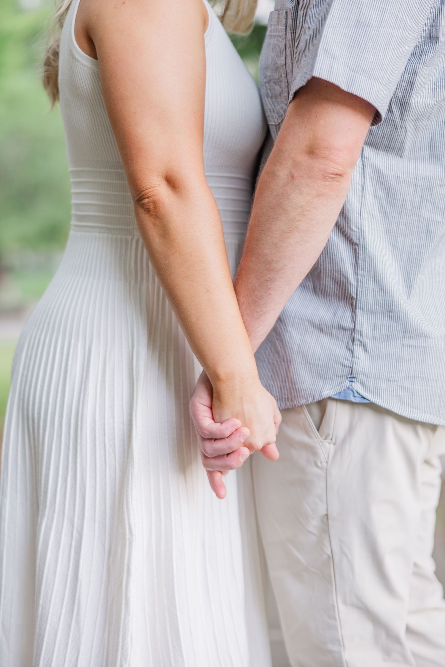 A couple holds hands tenderly while wearing casual wedding attire, with a white pleated dress and gray shirt visible.