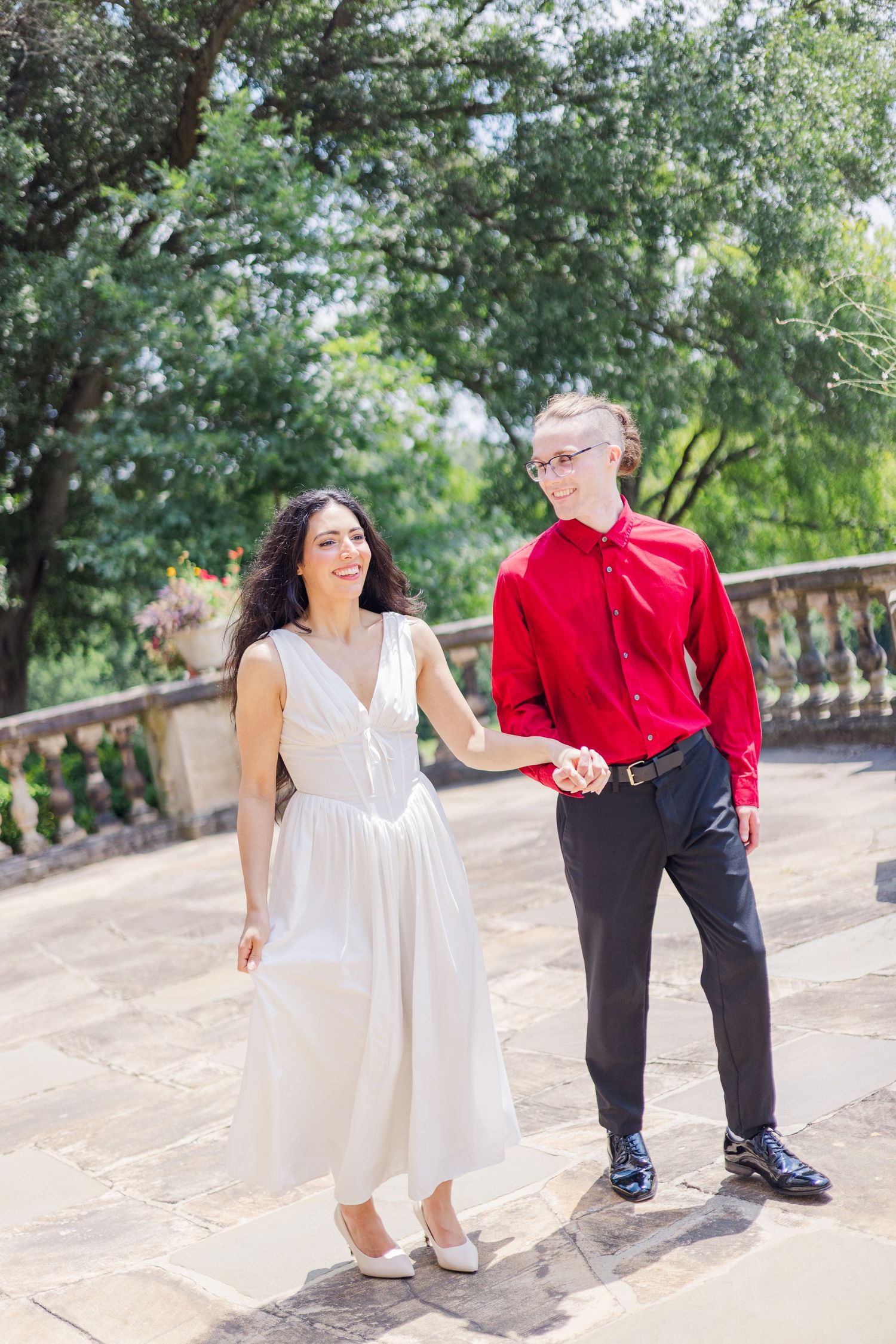 A couple in white dress and red shirt poses playfully on stone steps in a garden setting.