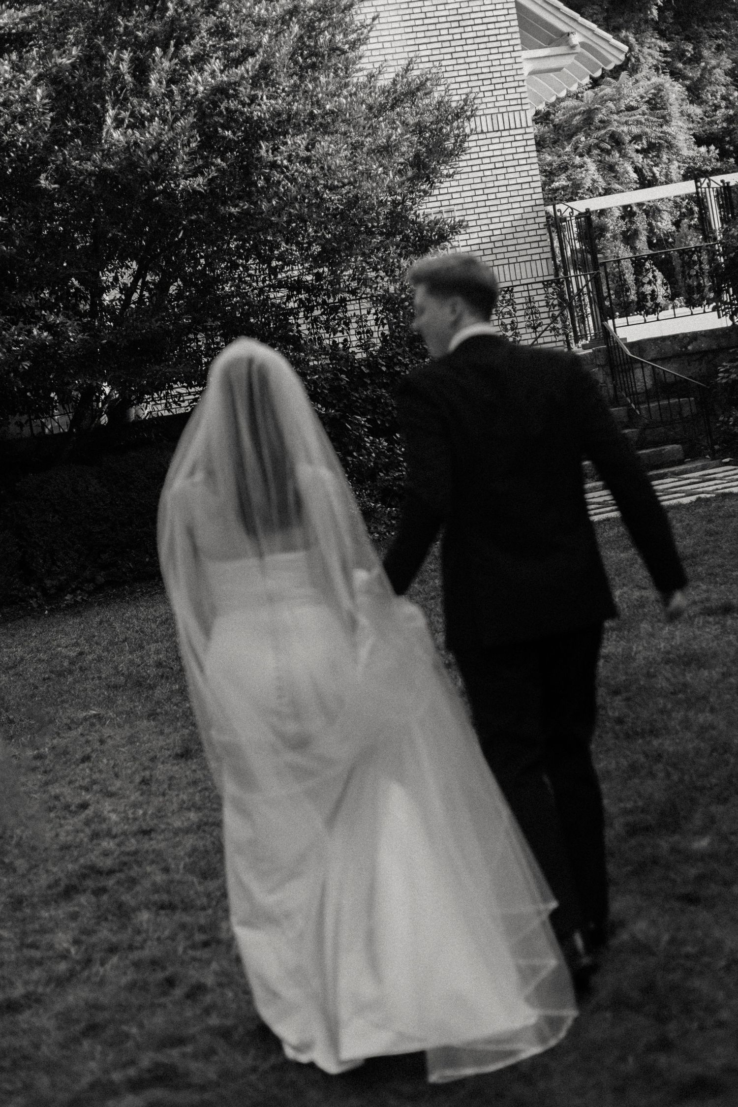 Black and white artistic photo of a couple walking through a garden with flowing veil.