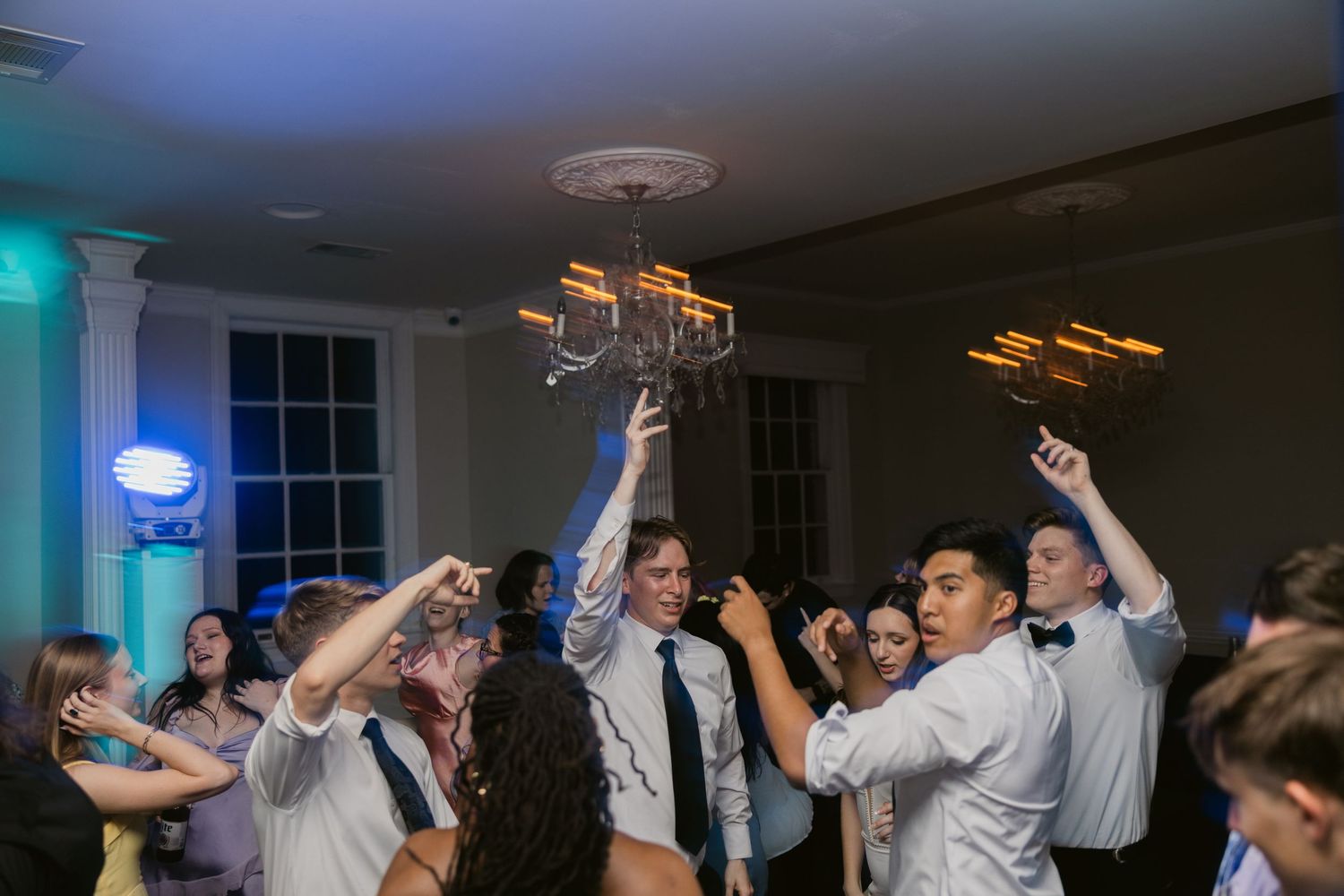Wedding party celebrating on dance floor with raised hands under decorative lighting.