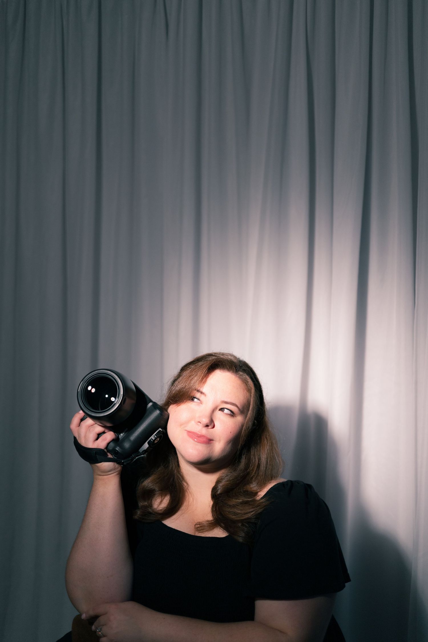 Woman in black top holding a professional DSLR camera, posing confidently against a gray curtain backdrop.