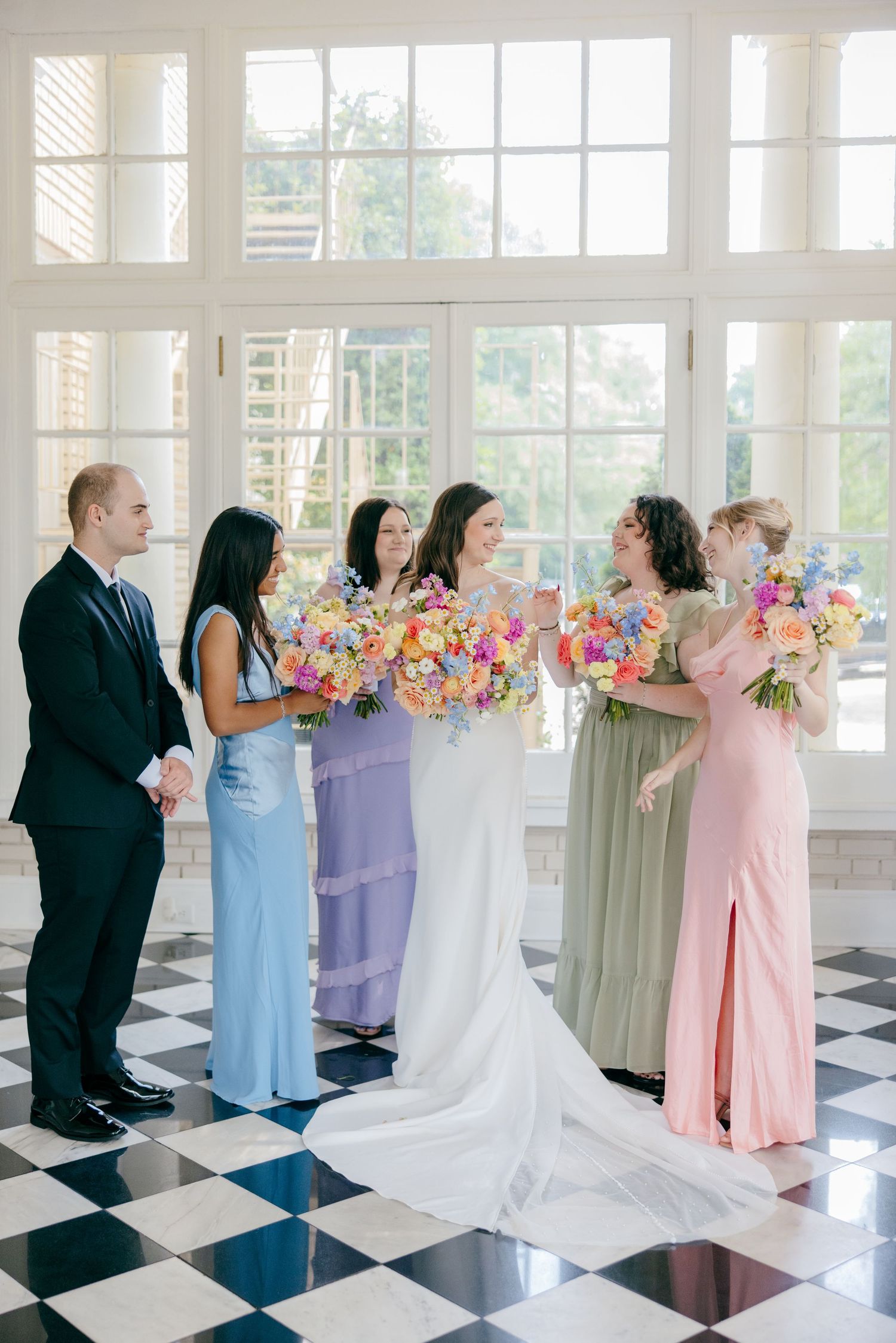 Wedding party lineup holding colorful bouquets in a bright venue with black and white tile flooring.