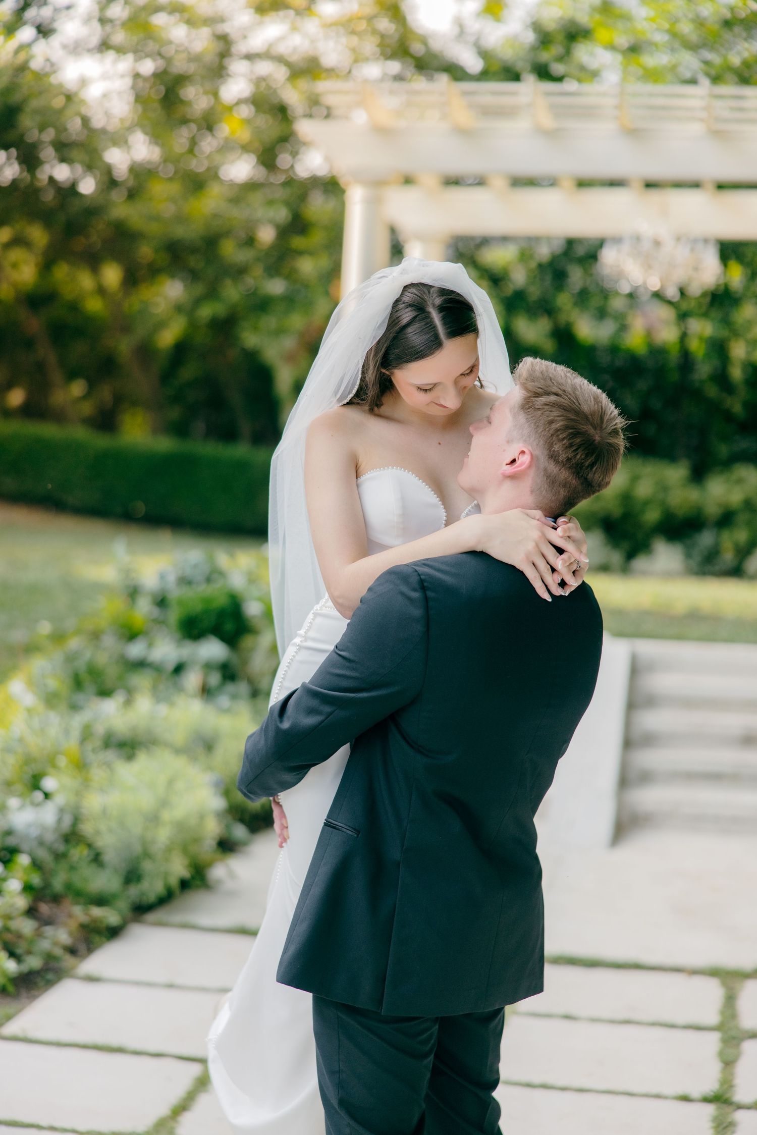 Tender embrace between newlyweds in an outdoor garden setting with white pergola.