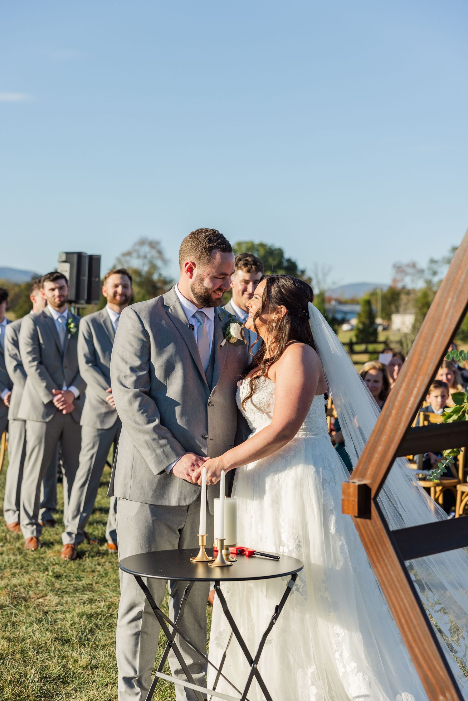 Wedding ceremony taking place outdoors with grey-suited groomsmen and white-dressed attendants.