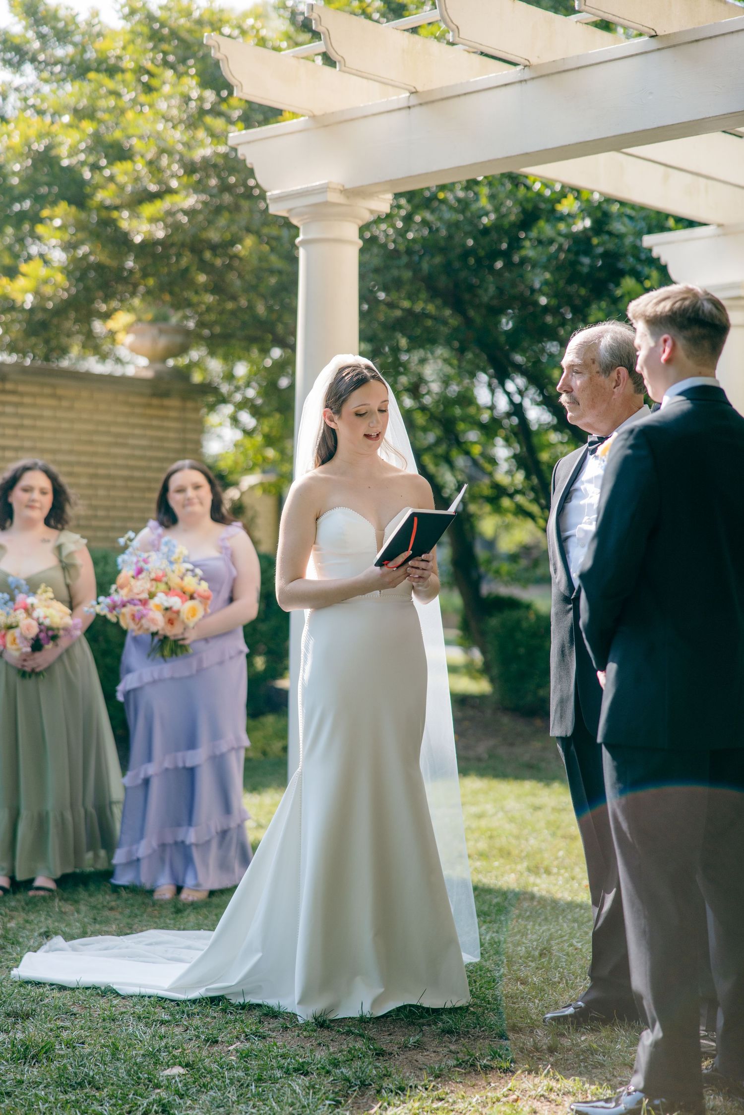 Outdoor wedding ceremony under a white columned pergola with pastel-dressed attendants.