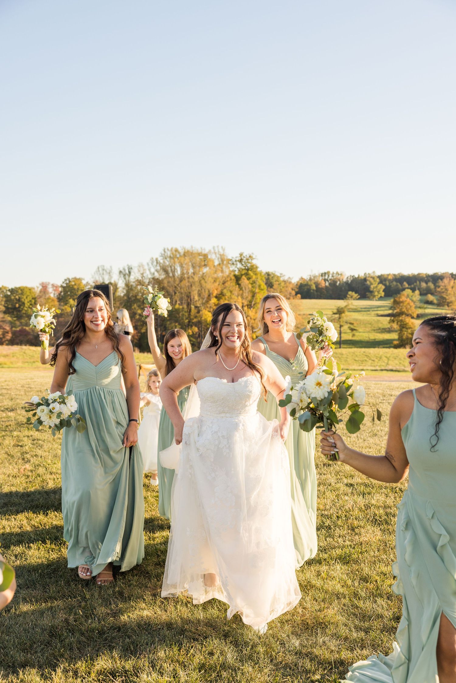 Bridal party in mint green dresses walking and laughing together in a sunny field during golden hour.