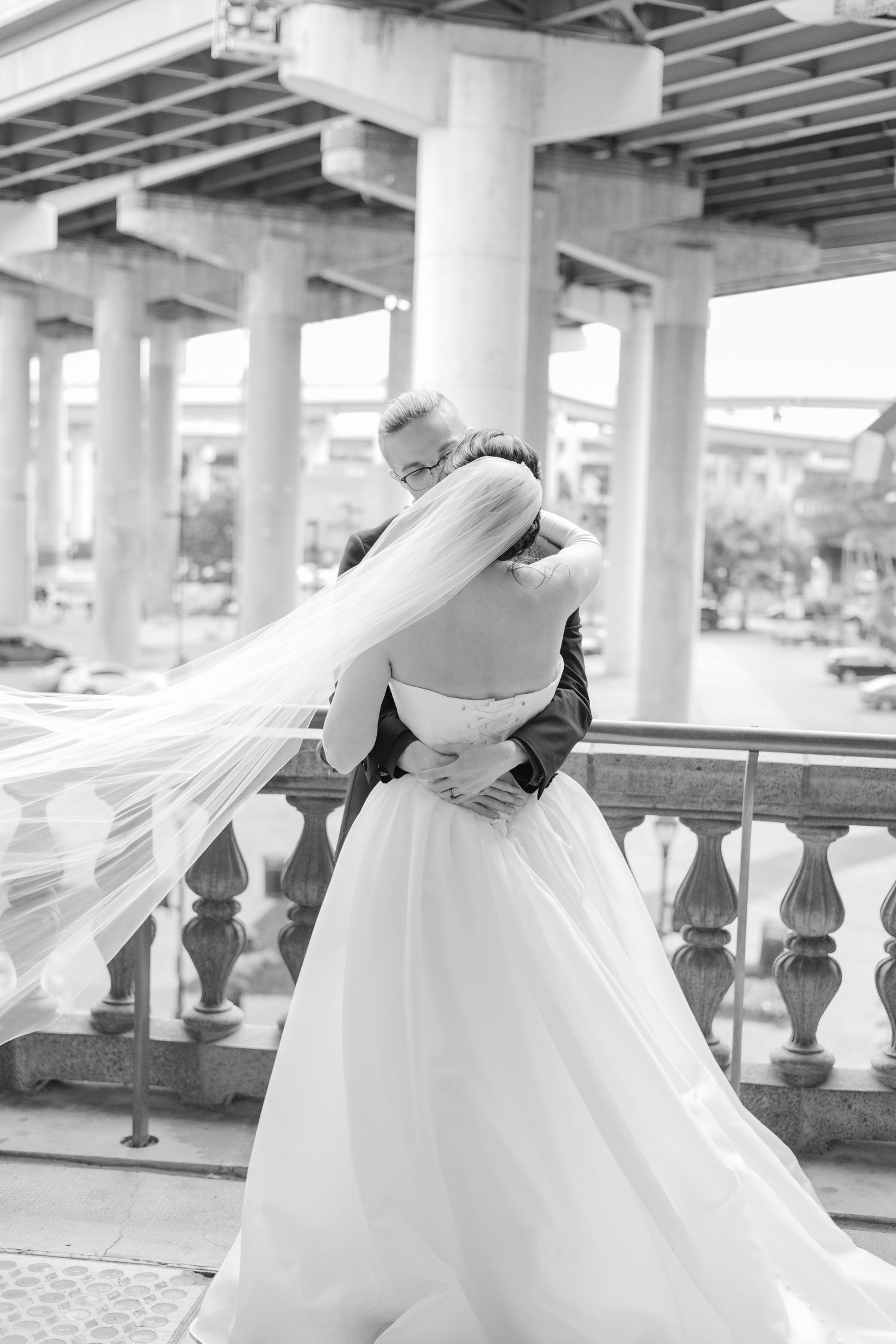Black and white wedding photo showing embrace under concrete pillars with flowing veil in romantic moment.