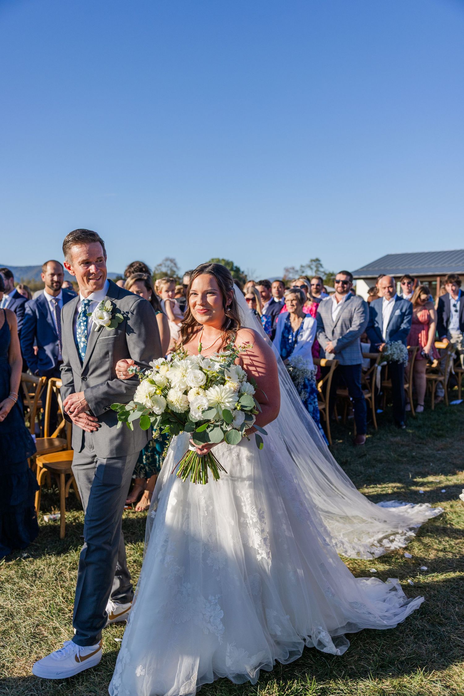 An outdoor wedding ceremony takes place on a grassy field with guests watching the celebration.