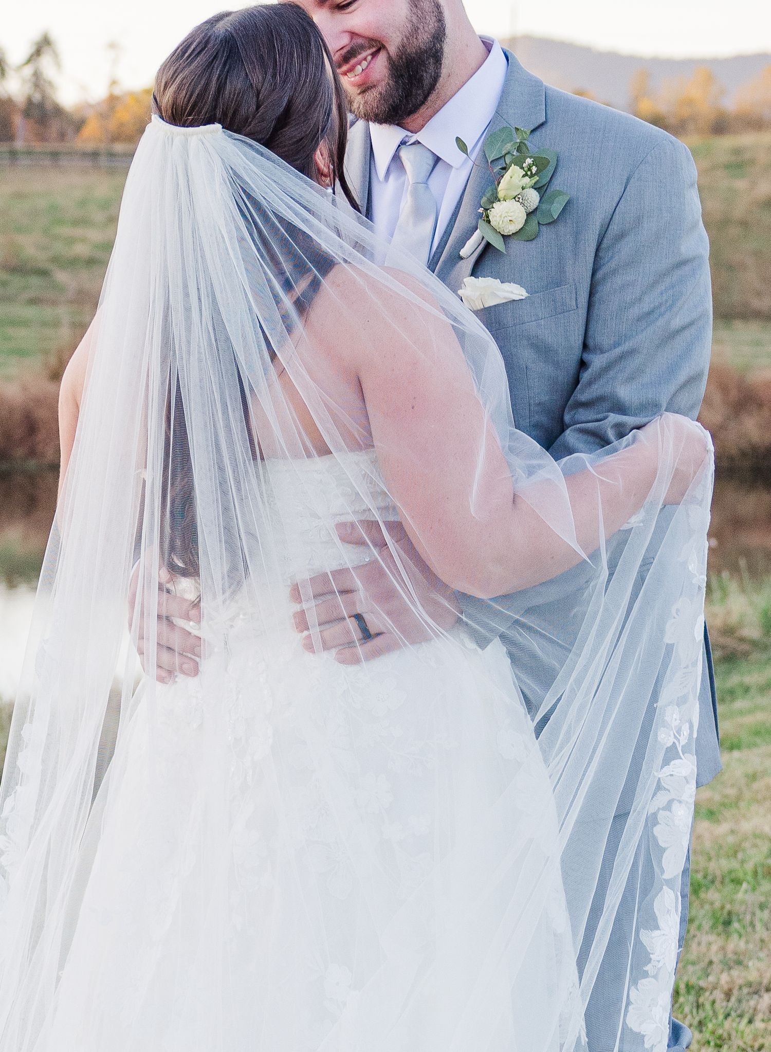 Romantic wedding couple embraces outdoors with flowing veil creating ethereal effect in soft autumn light.