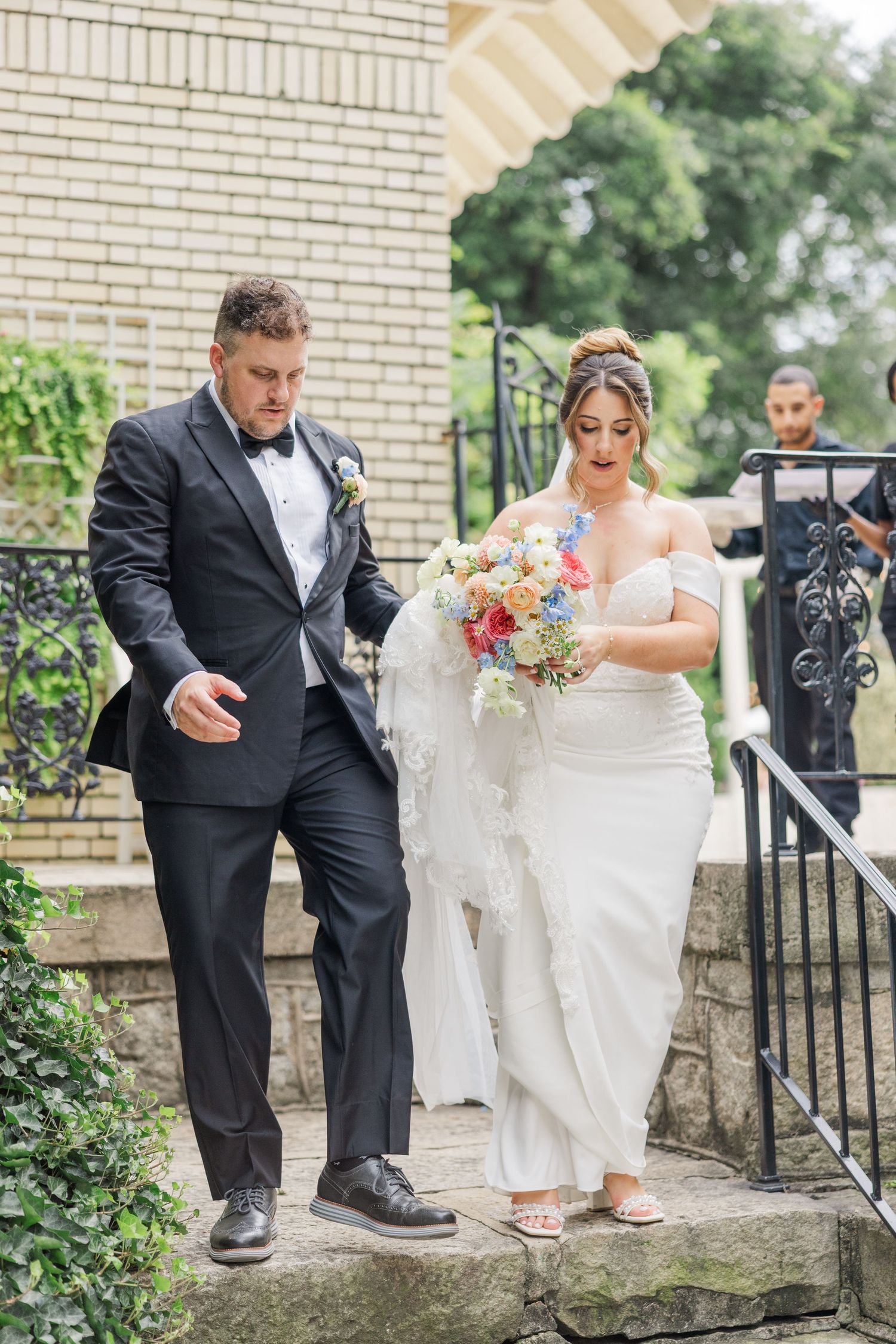 Wedding couple navigates stone steps together in formal attire with brick wall and railing details.