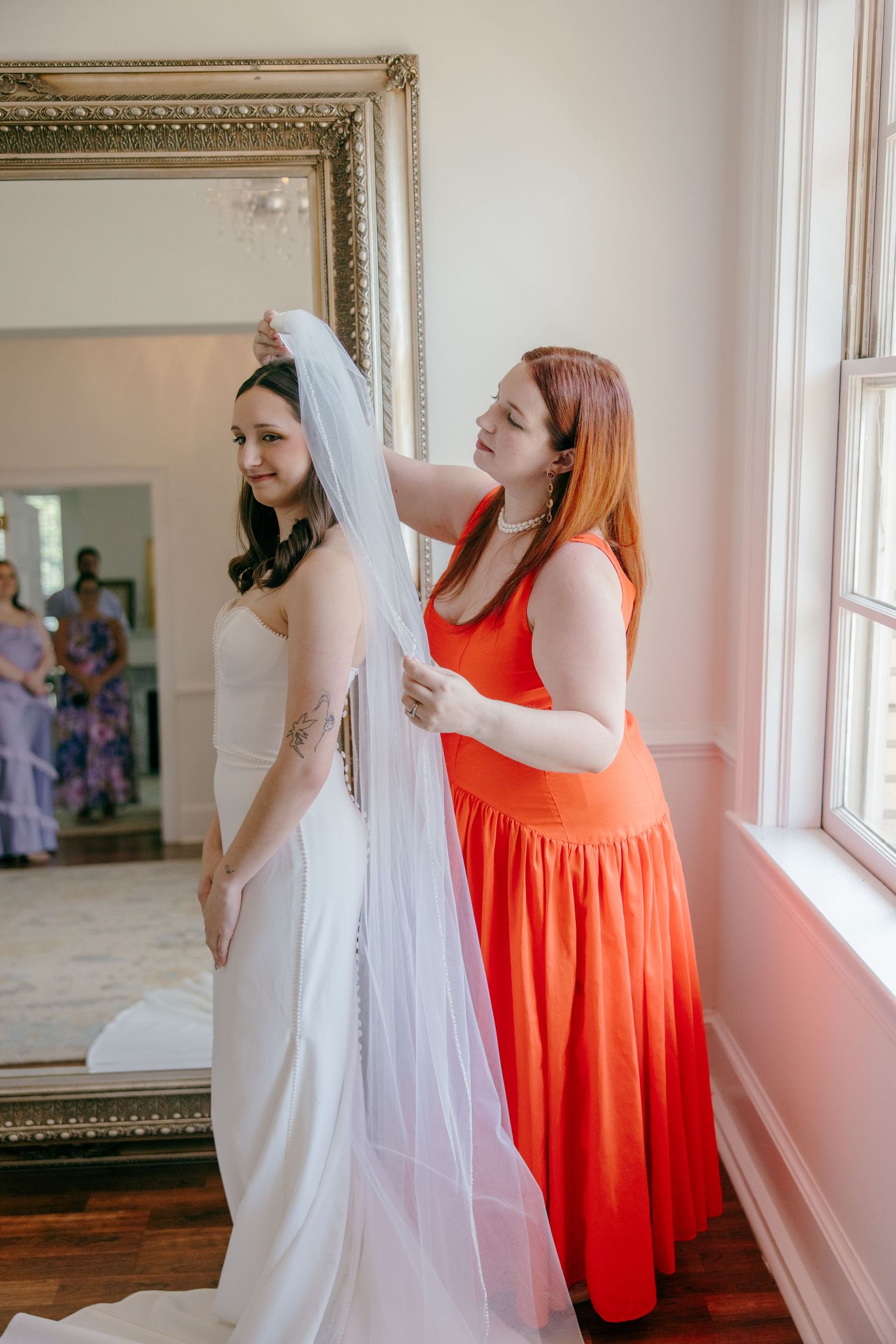 A wedding preparation scene with a flowing white veil and coral orange bridesmaid dress in a sunlit room.