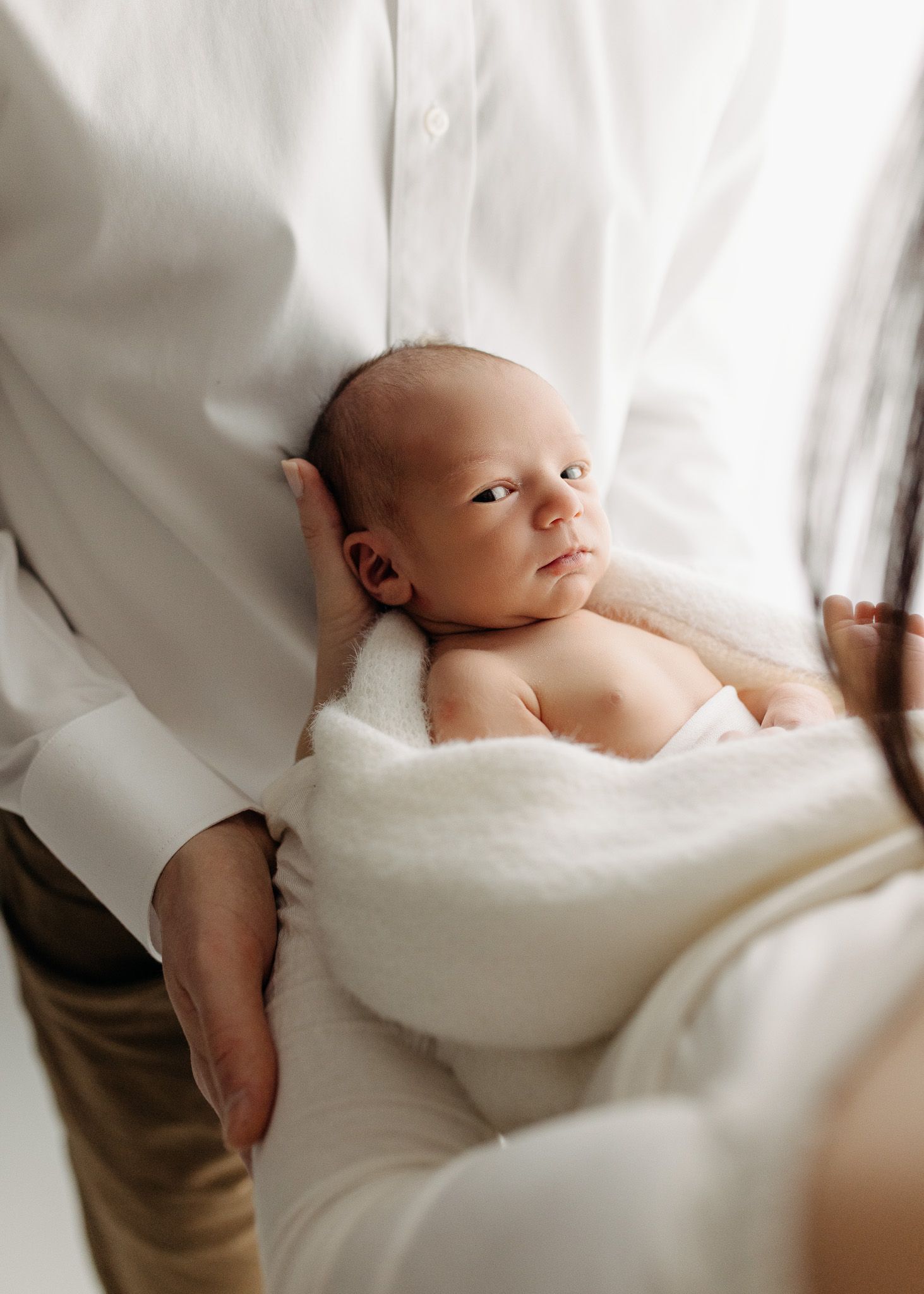 Newborn baby cradled in white blanket against bright background.
