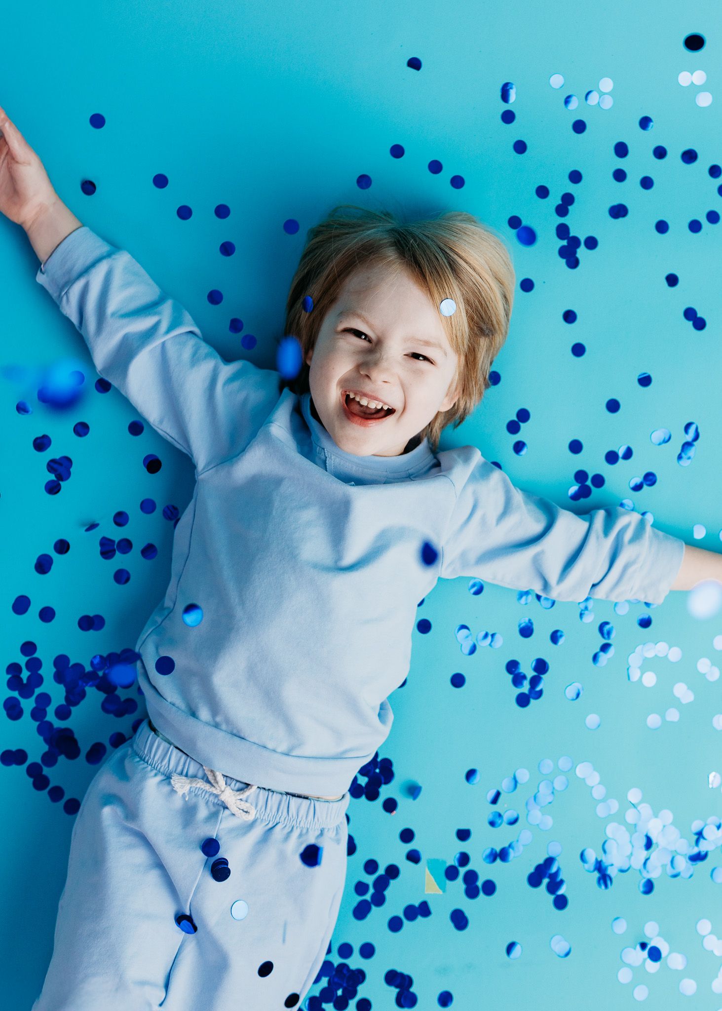 Young child lying on blue background with scattered confetti dots showing pure joy and excitement.