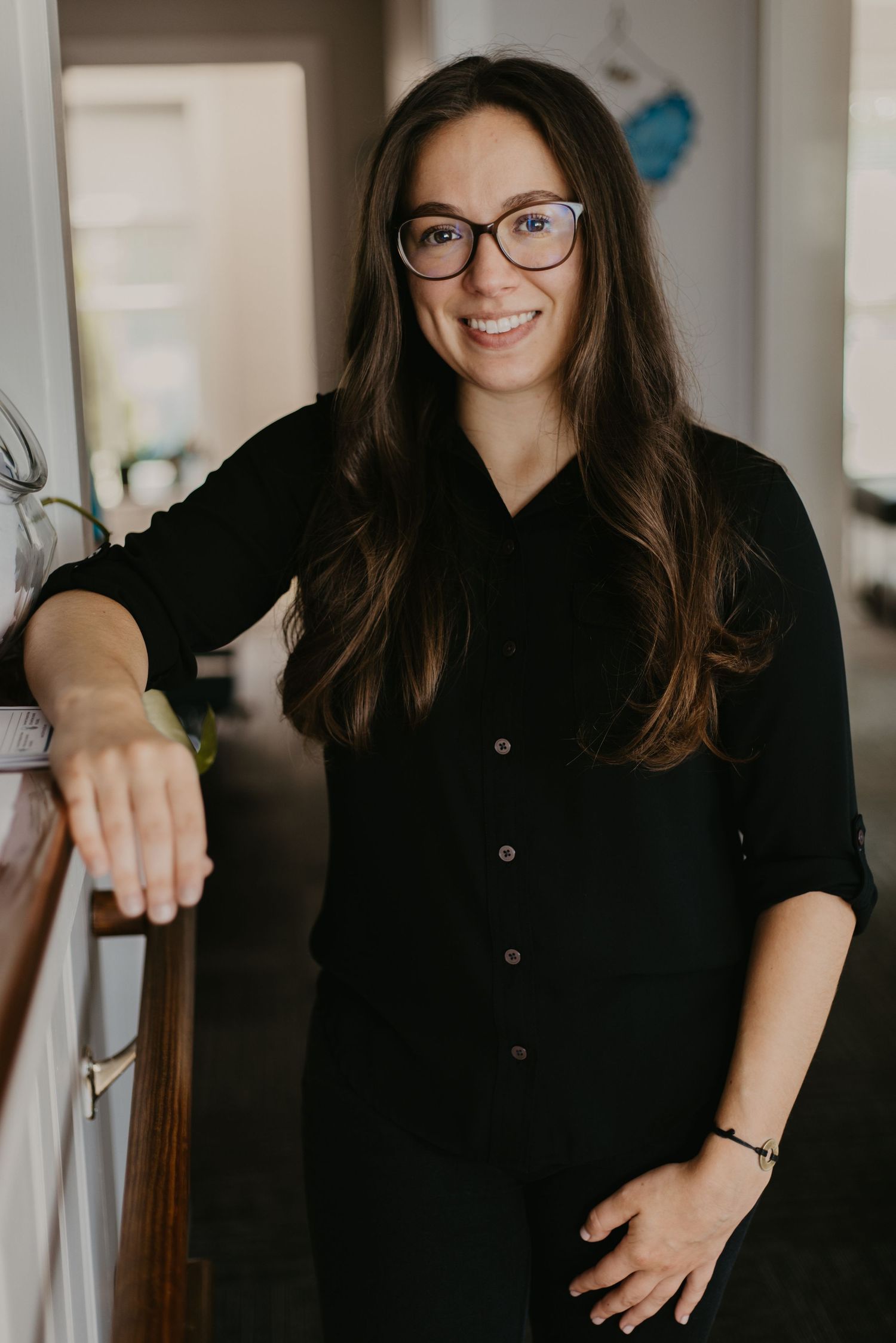 Smiling woman with glasses and long brown hair wearing a black outfit, leaning on a railing indoors.