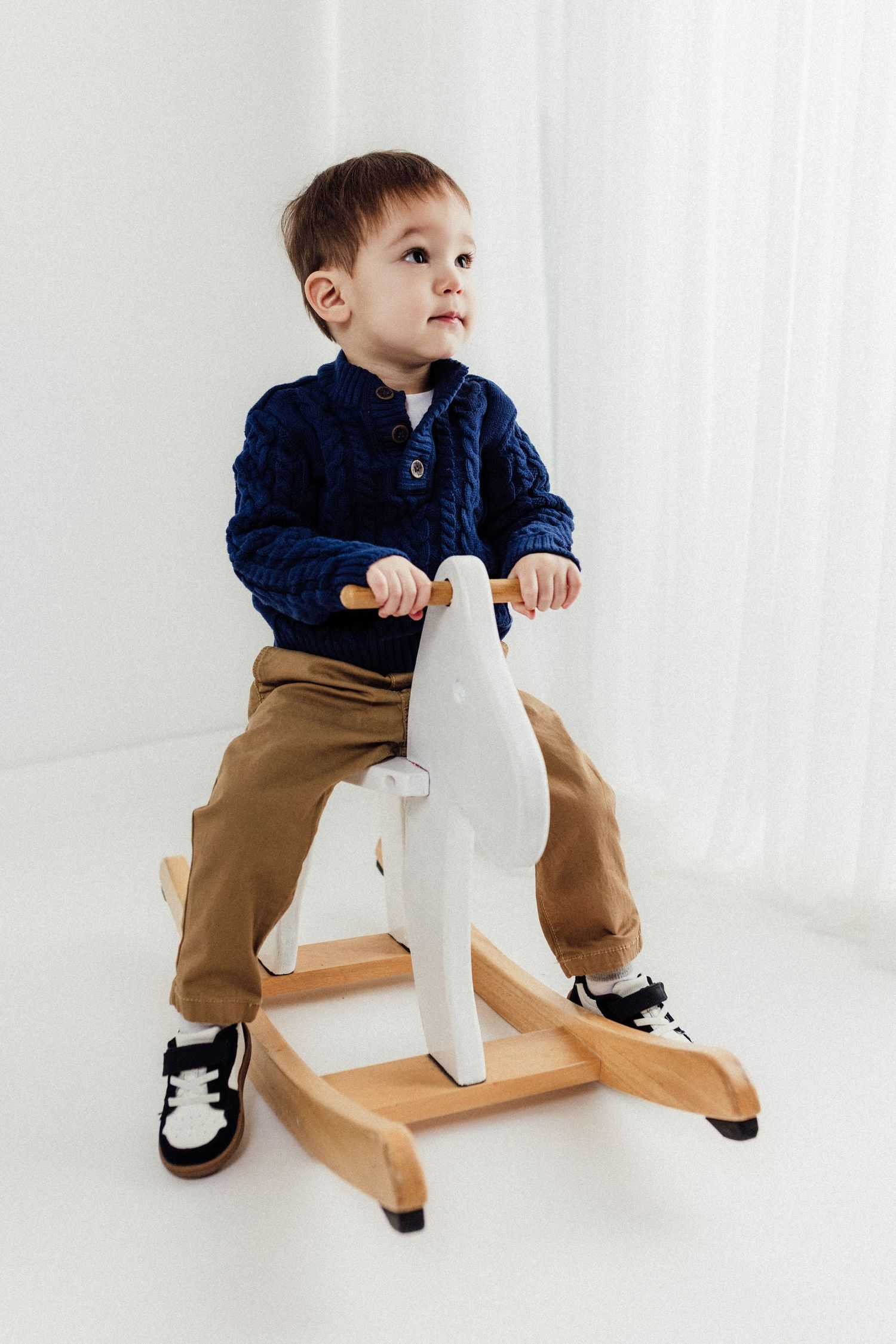 Child riding wooden rocking horse toy wearing navy jacket and khaki pants.