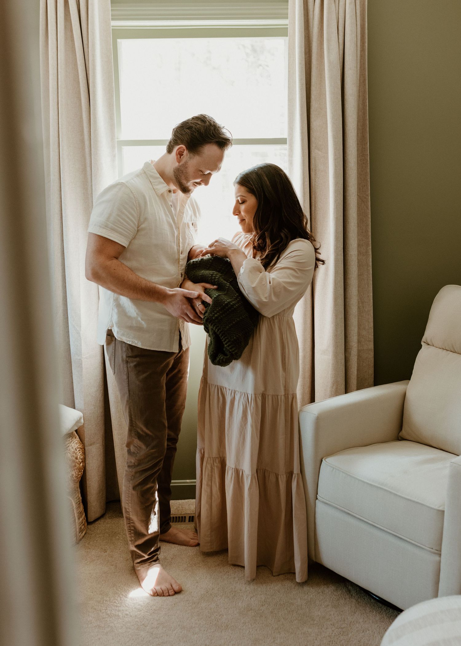 New parents stand by a sunlit window, lovingly gazing at their newborn baby wrapped in a green blanket.