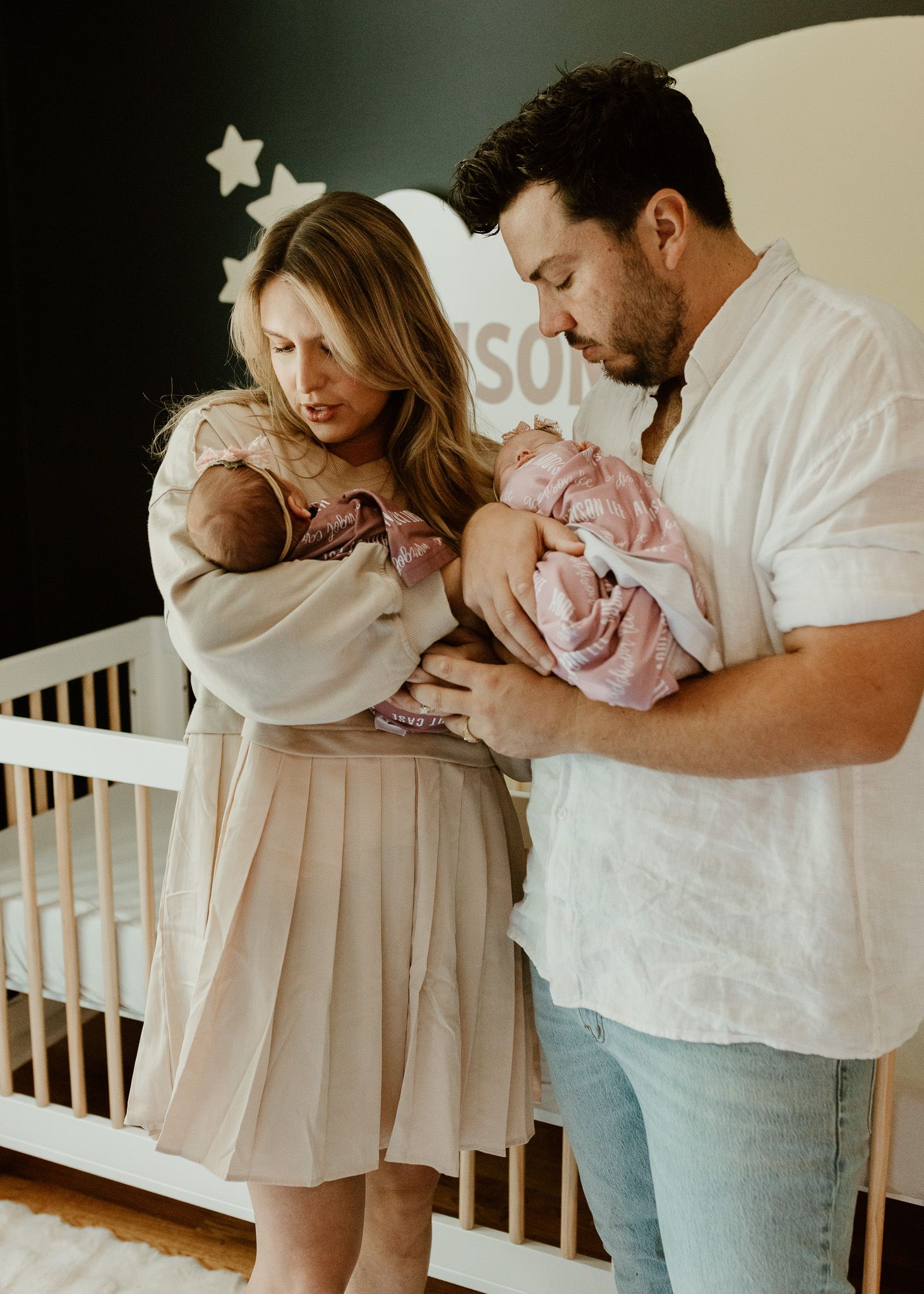 Parents in a nursery holding and gazing lovingly at their newborn twins wrapped in pink blankets.