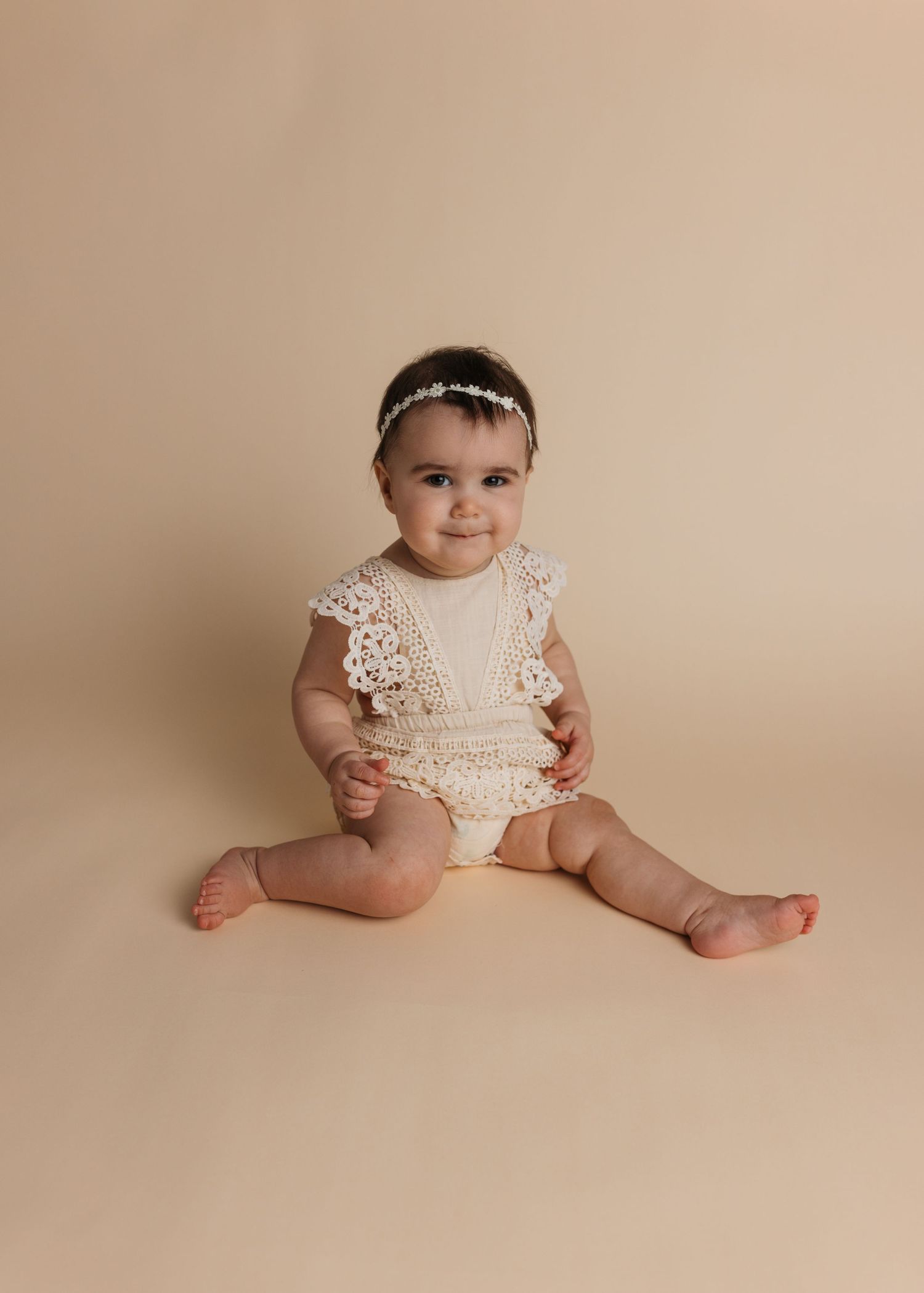 Baby wearing lace romper sitting on beige backdrop during professional photoshoot.
