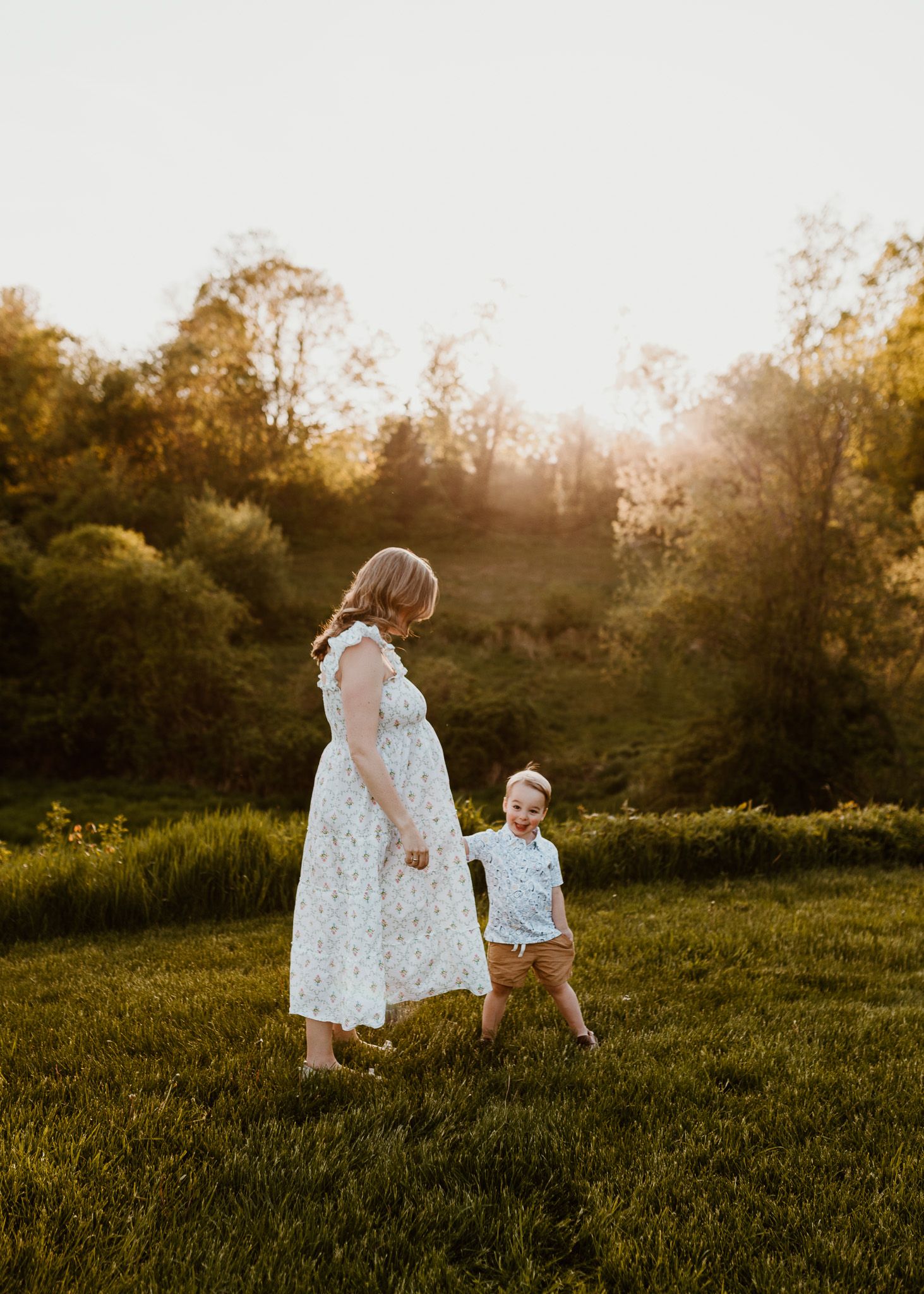 Pregnant woman in floral dress walks with toddler boy in sunny green meadow at golden hour.