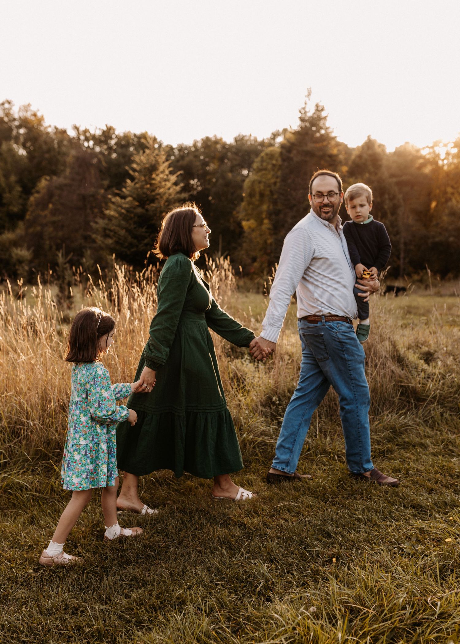 Family of four walking in golden hour field, parents holding hands with young children, surrounded by tall grass and trees.
