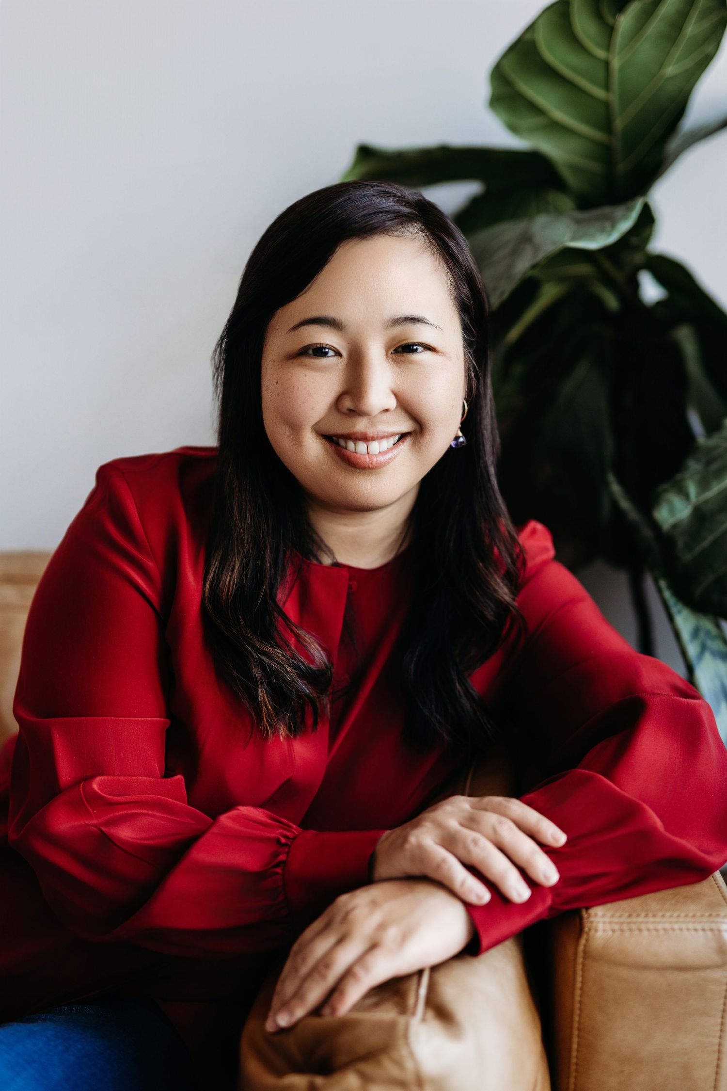 Smiling Asian woman in red blouse seated on leather sofa with tropical plant in bright modern interior.