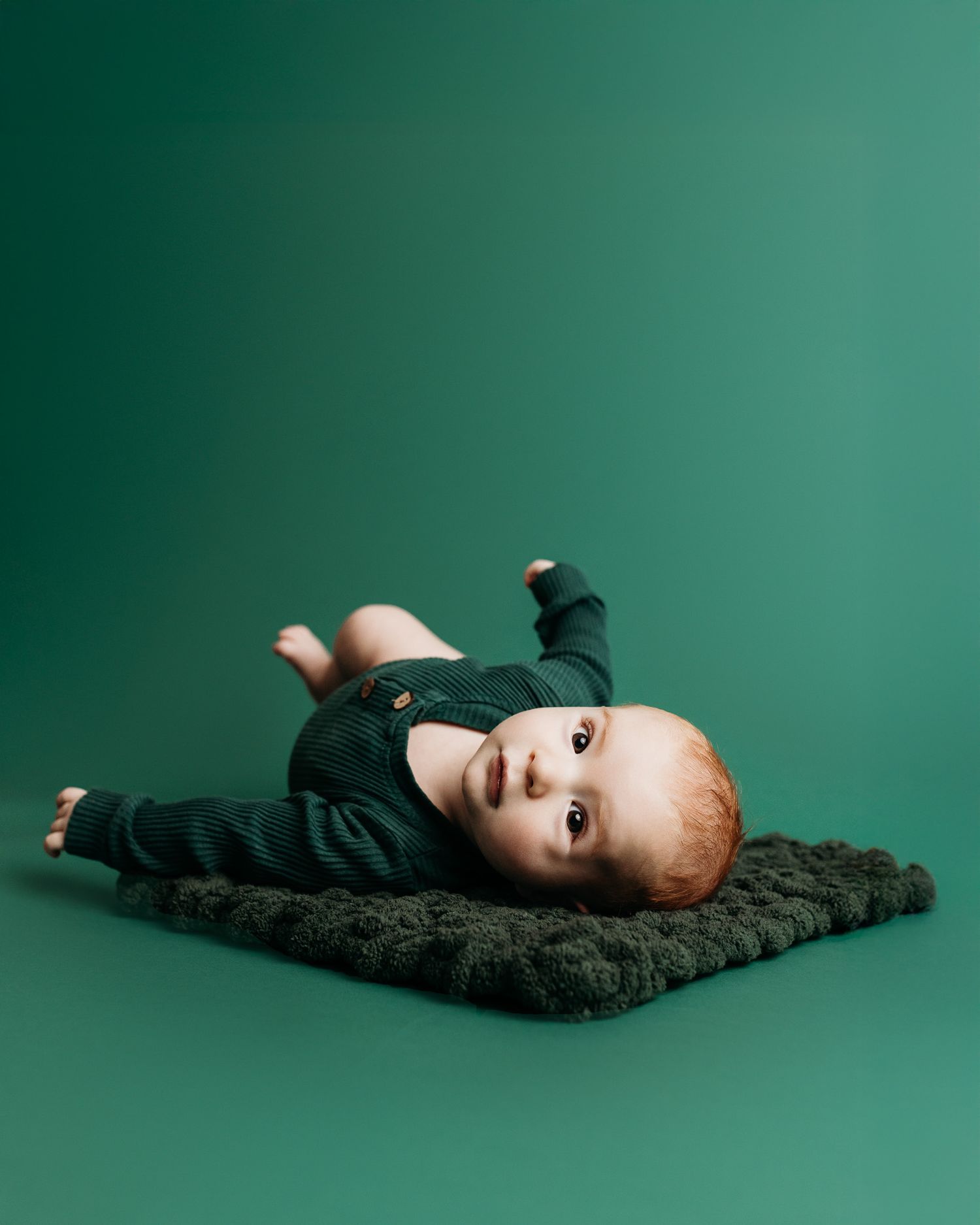 Baby in dark outfit lying on knit blanket against green background.