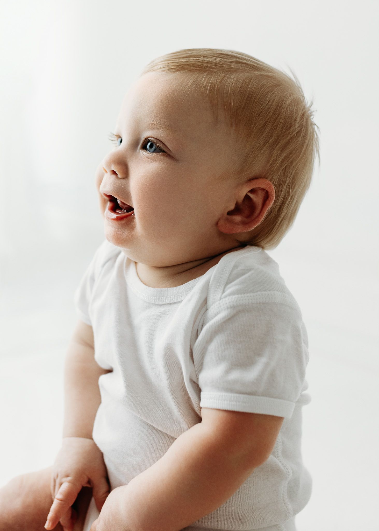 A happy blonde toddler in a white onesie smiles joyfully against a bright white background.