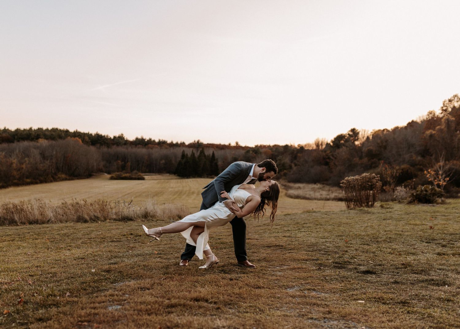 Romantic couple sharing a dip kiss in an open field at golden hour with trees in the background.