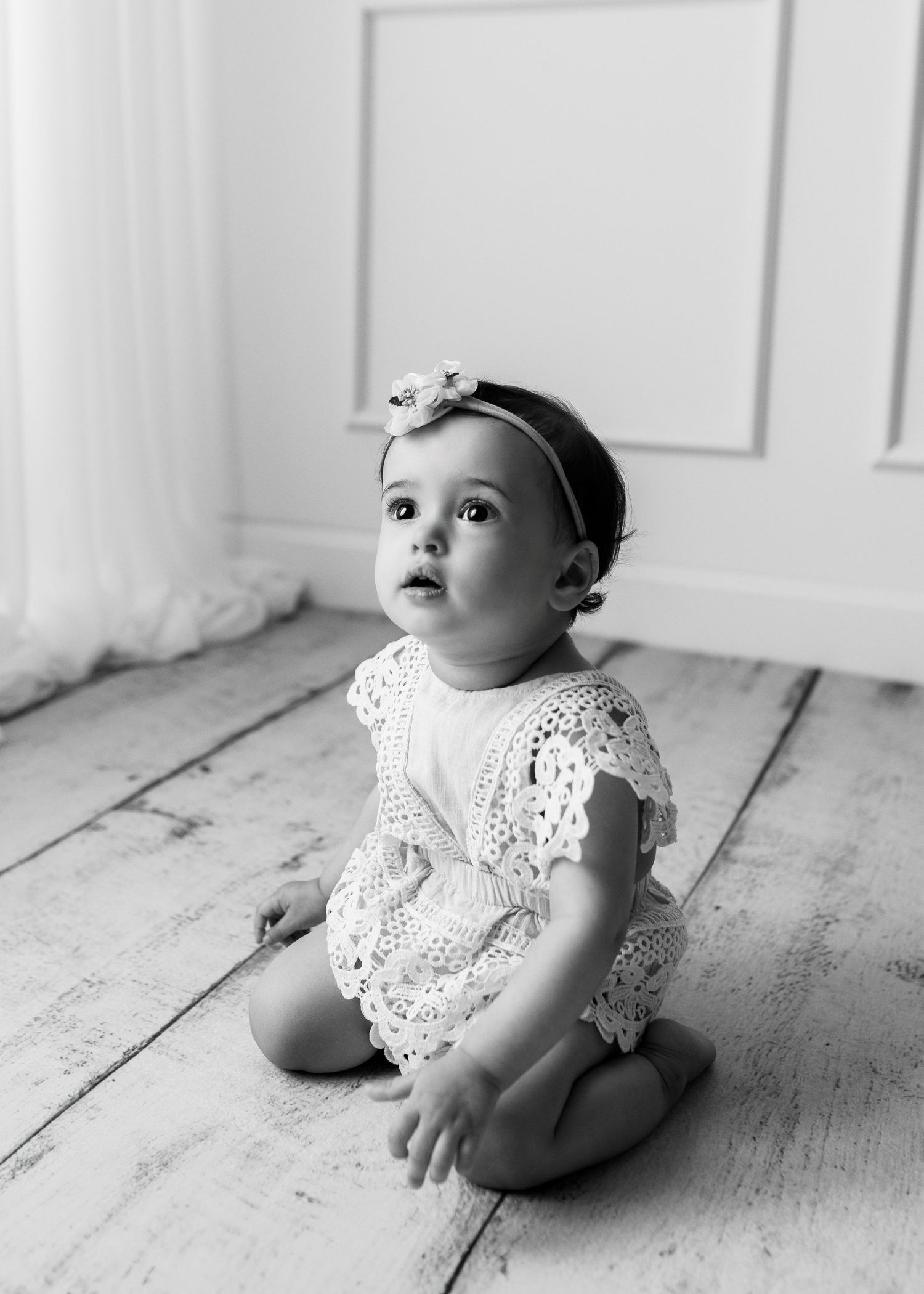 Black and white photo of a baby girl in lace romper and bow headband sitting on wooden floor looking up.