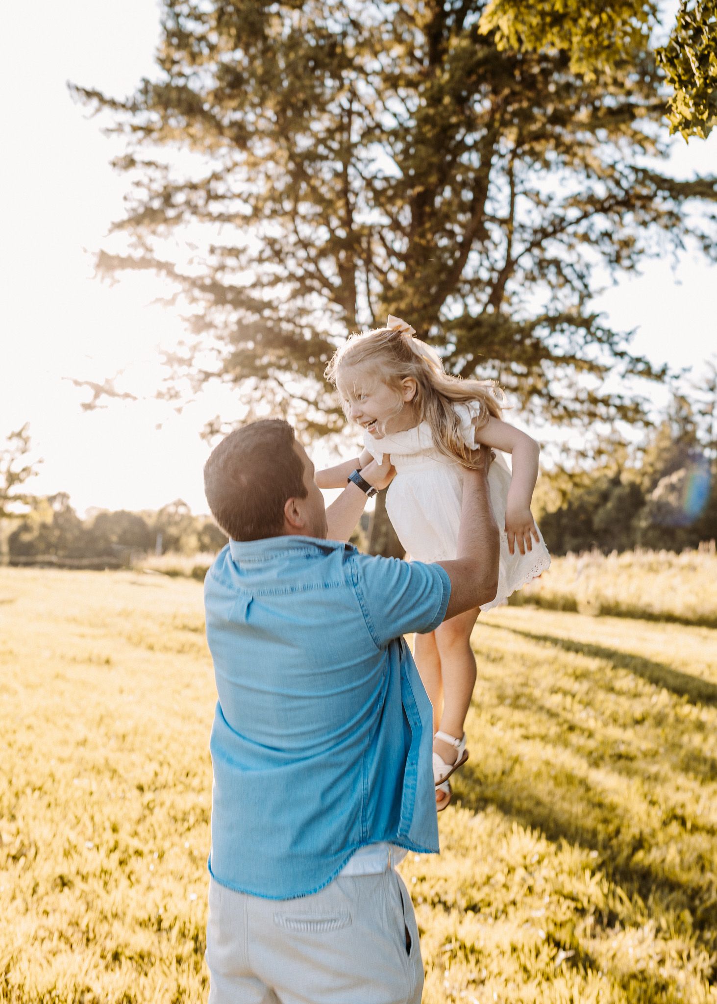 A father lifts his laughing young daughter in a sunlit park, both wearing light clothing amid golden hour greenery.