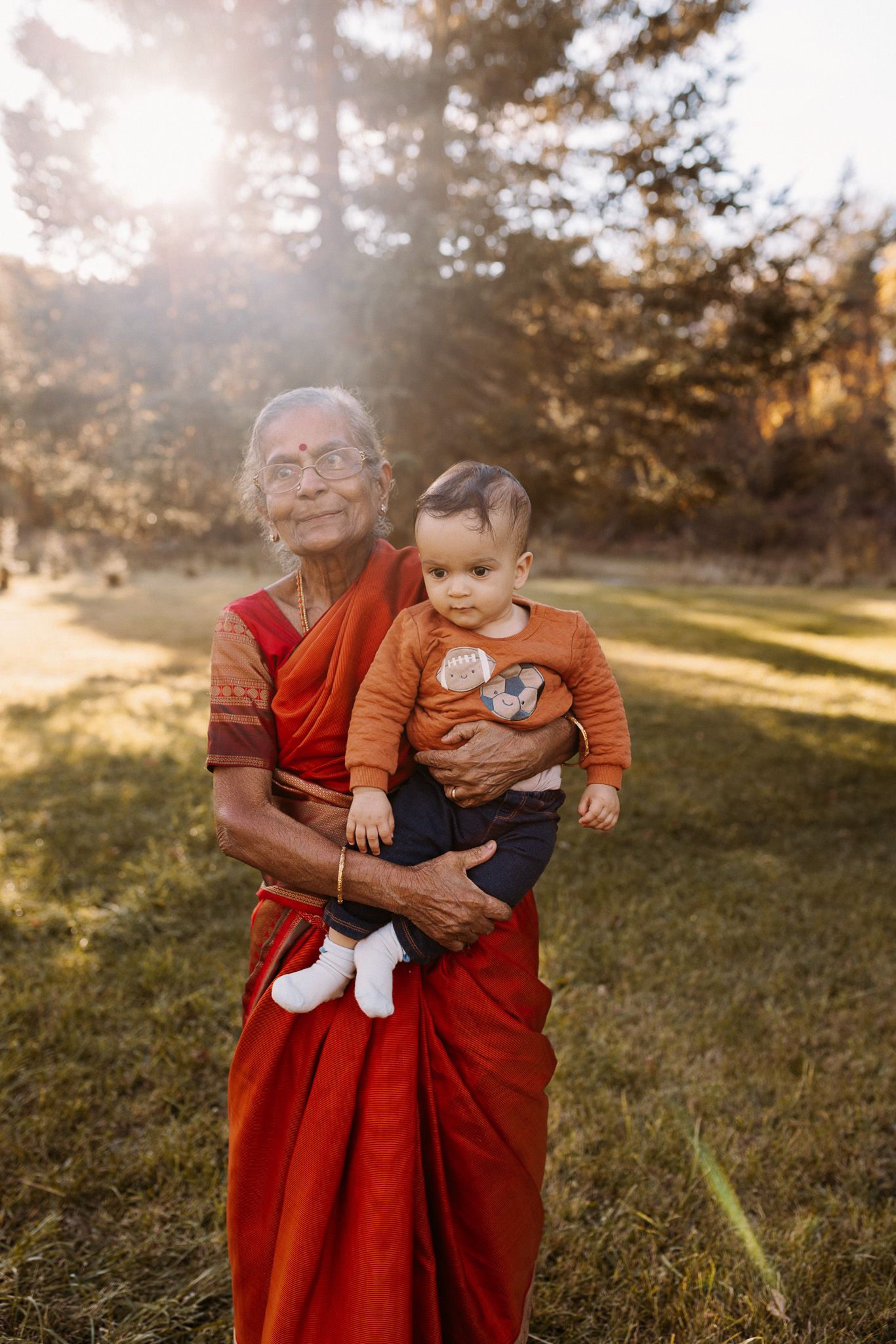 Elderly Indian woman in red saree holds a baby outdoors in golden sunlight with trees in background.