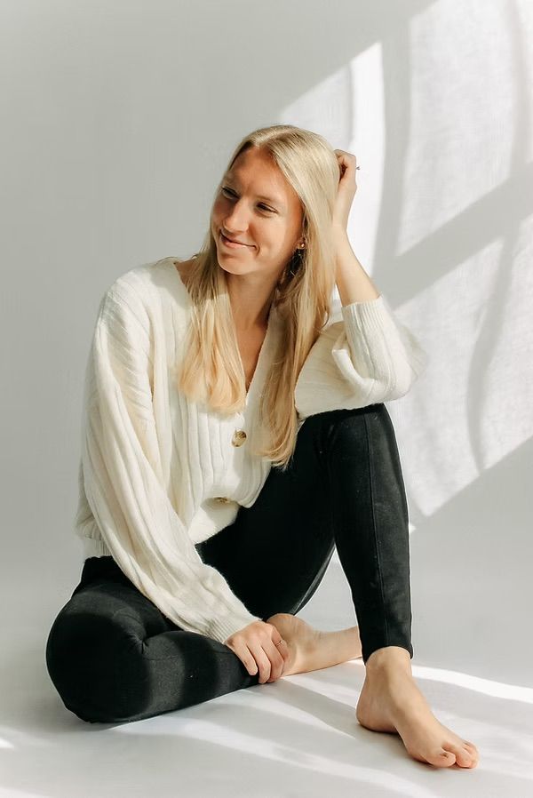 Woman in white sweater and black pants sitting casually against white wall with natural lighting.