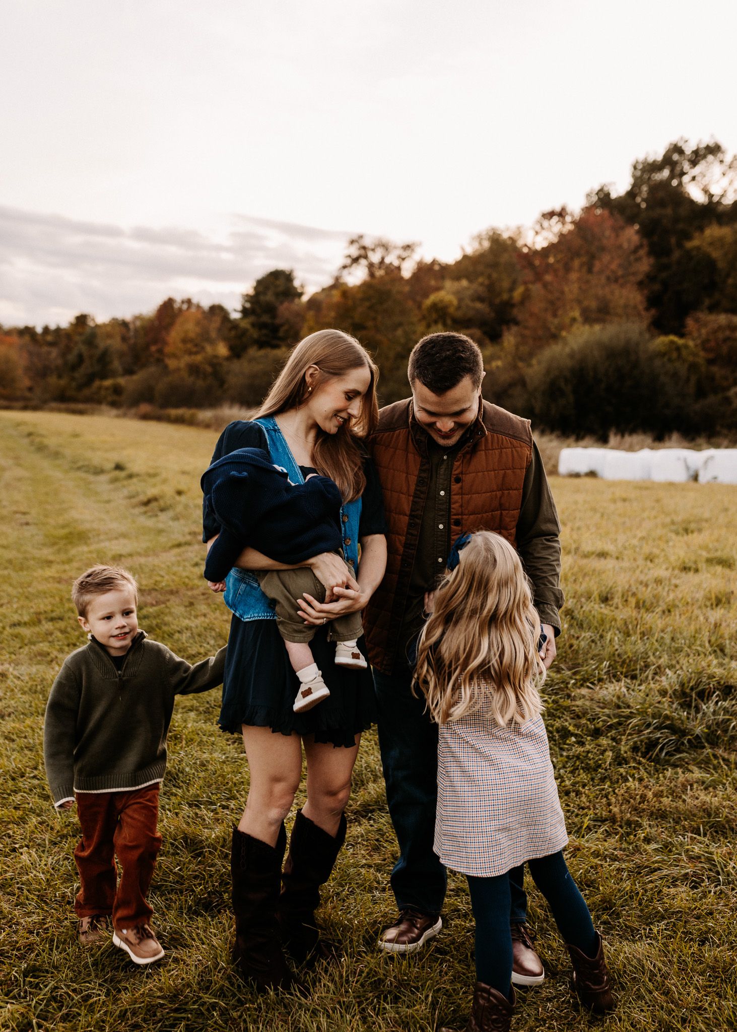 Family of five sharing a joyful moment in an autumn field, parents holding a baby while two children stand nearby.