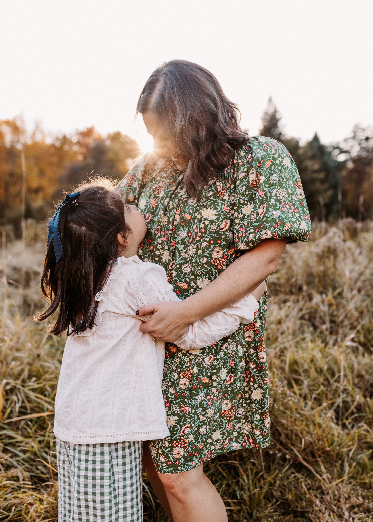Mother and young daughter embracing in golden sunset light in an autumn meadow, wearing floral and gingham outfits.