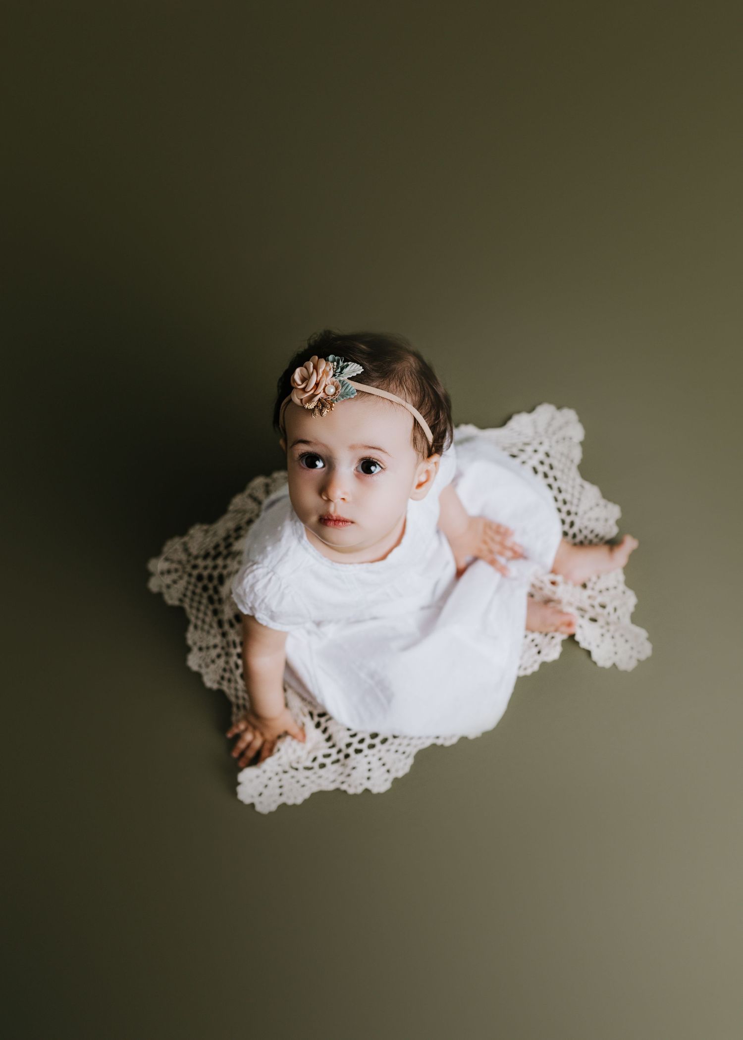 Overhead view of baby in white lace dress lying on dark background.